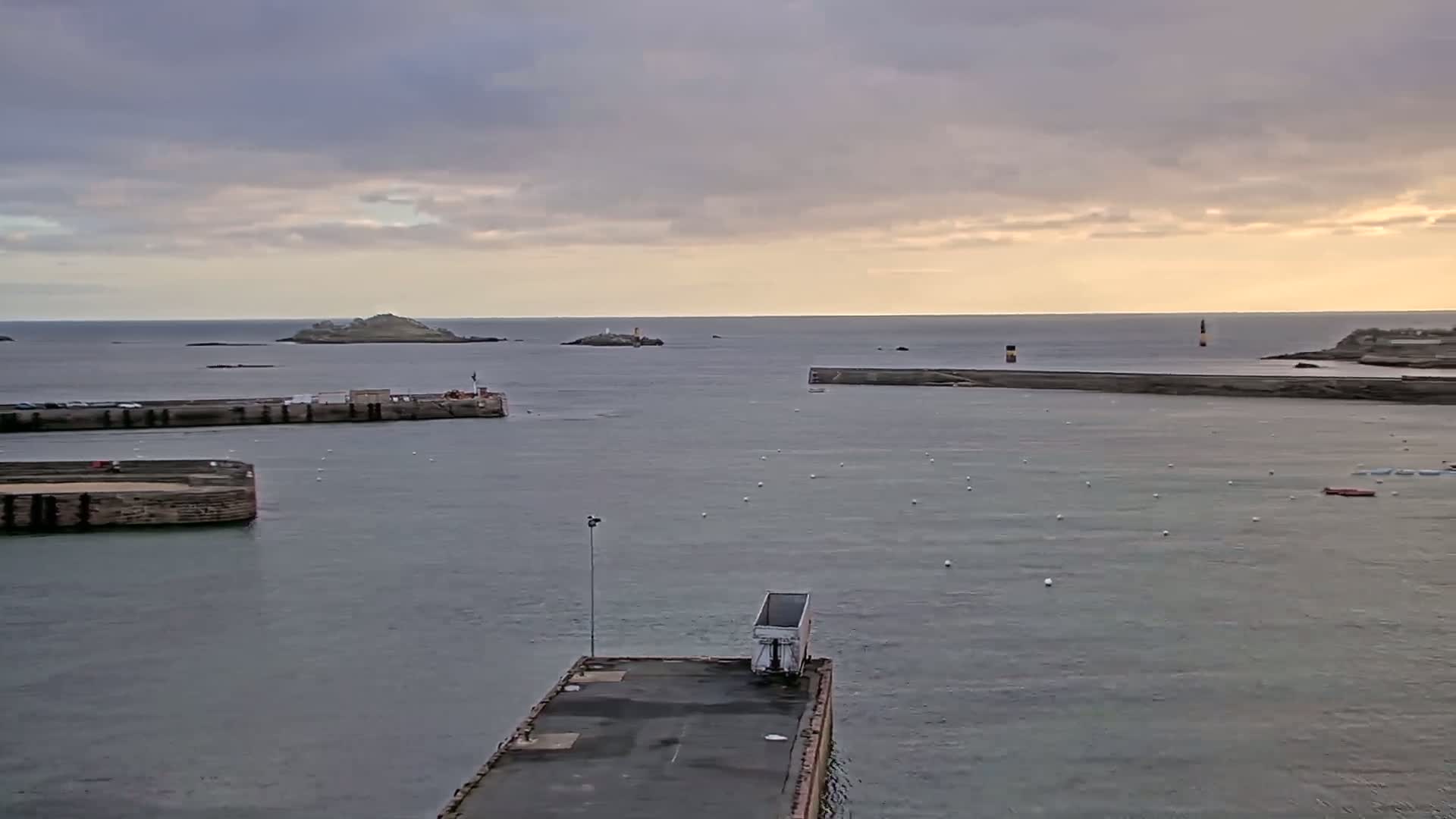 A harbor with calm water under an overcast sky shows several small islands and moored boats in the distance, with a lone truck on a pier in the foreground.
