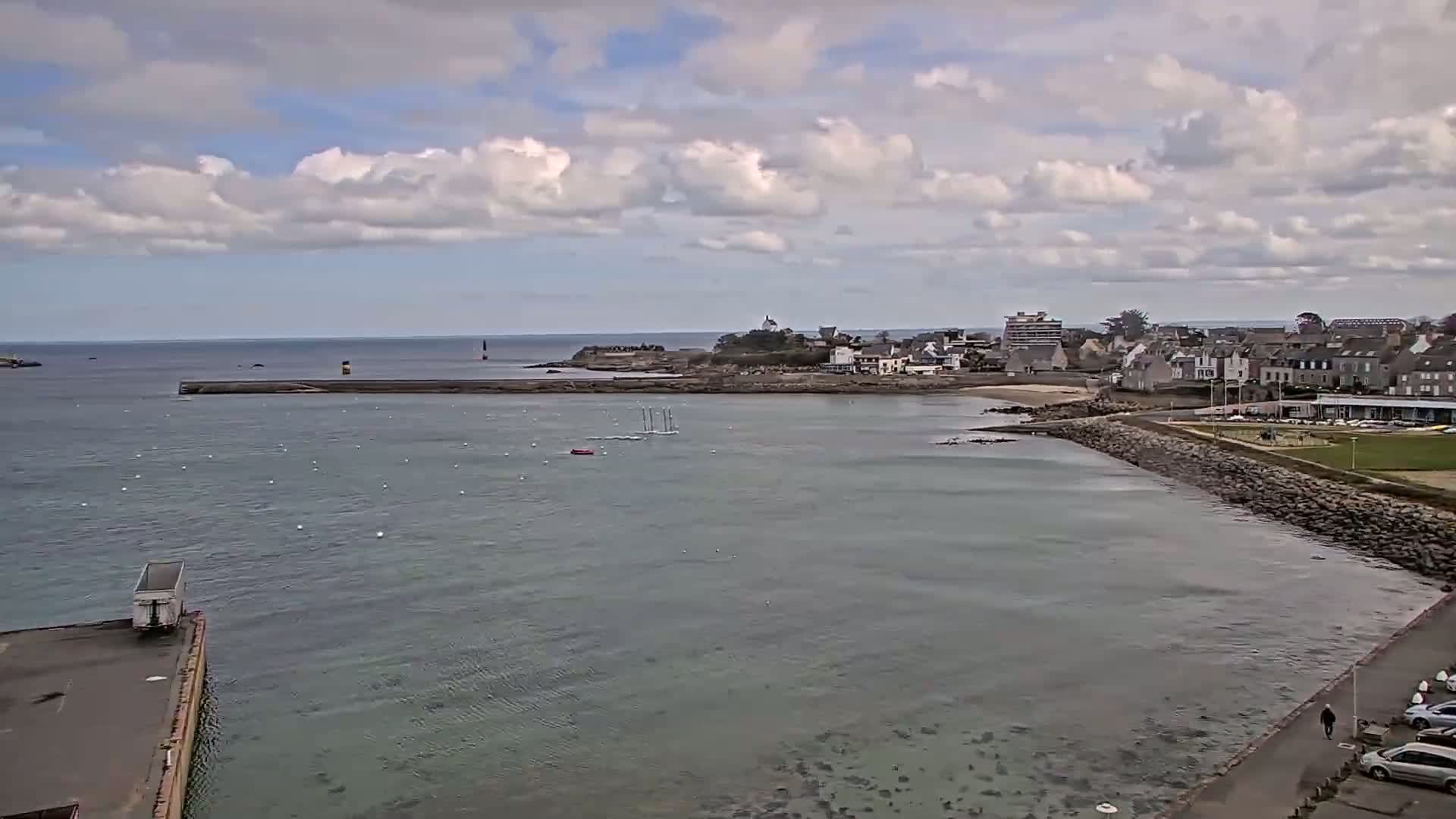 A partly cloudy day shows a coastal town with a calm bay containing several sailboats, a rocky shoreline, and a few parked cars.