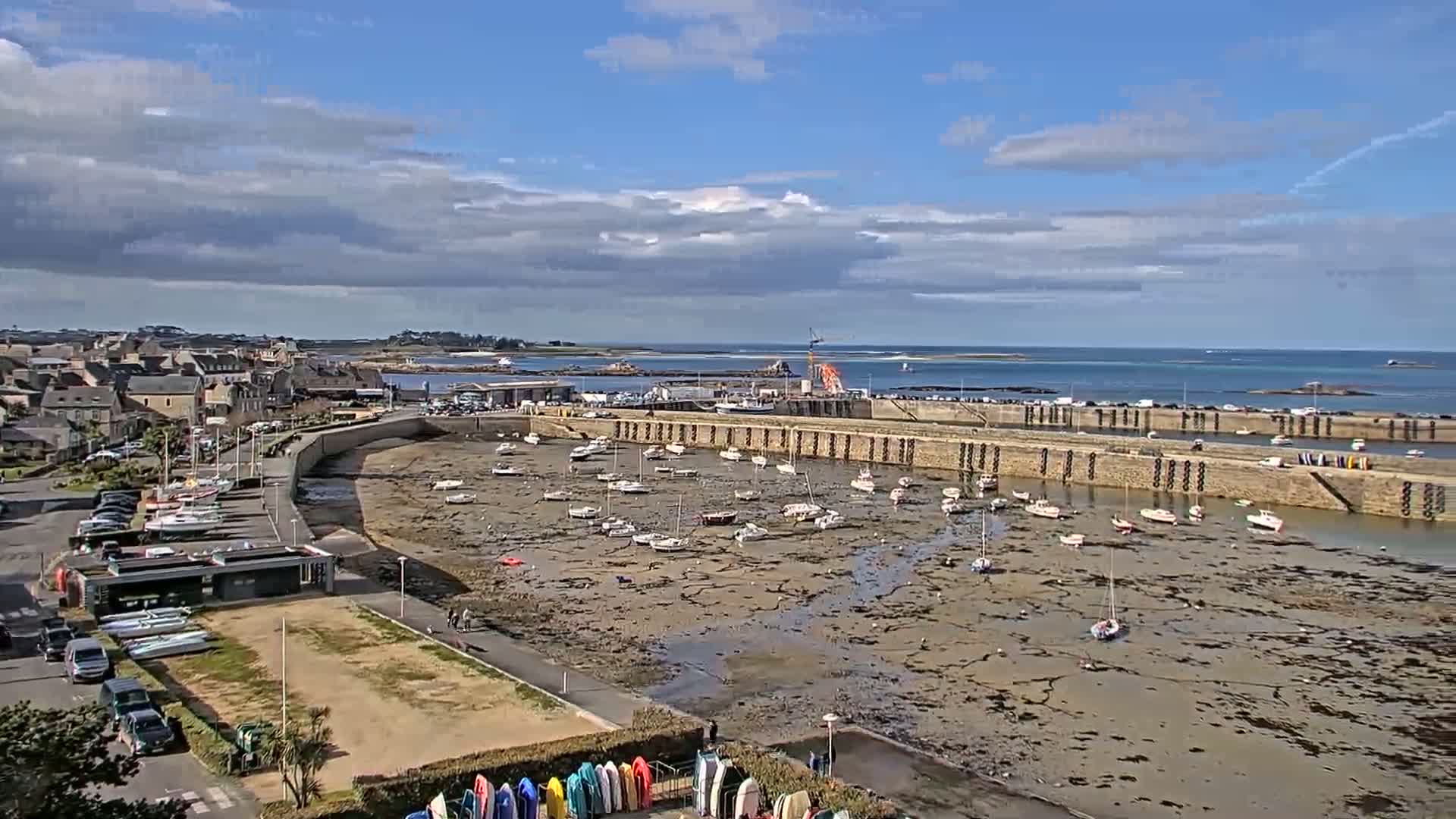 A partly cloudy day reveals a harbor with numerous small boats resting on exposed mudflats, adjacent to a town and a larger body of water beyond.