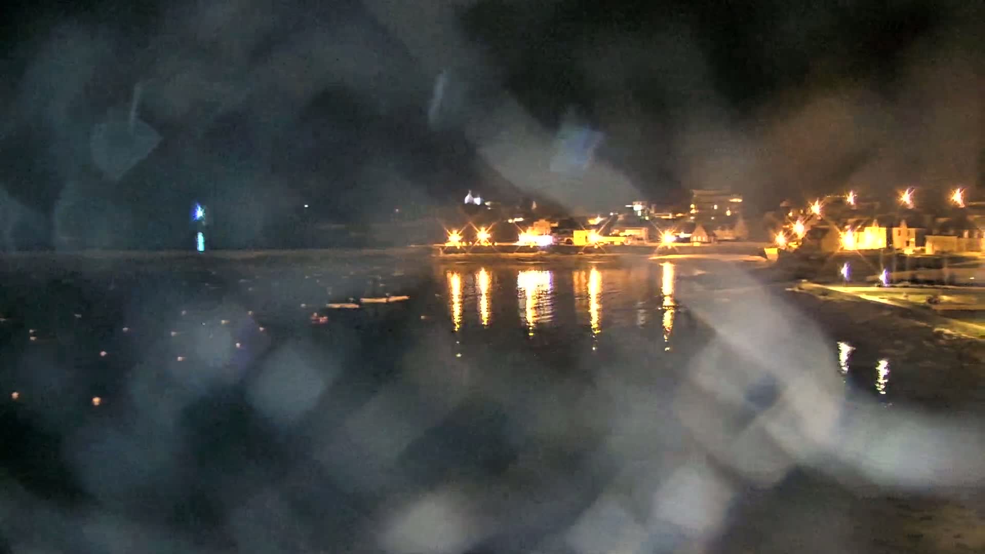 A nighttime view of a calm body of water reflecting the lights of a town across the shore, seen through rain-covered glass.
