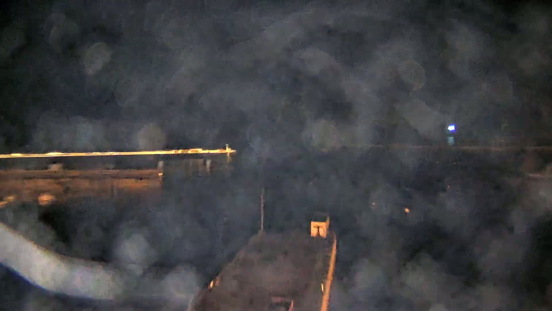 A blurry nighttime image shows a dark body of water with a distant line of lights and a small structure in the foreground, under a hazy or foggy sky.