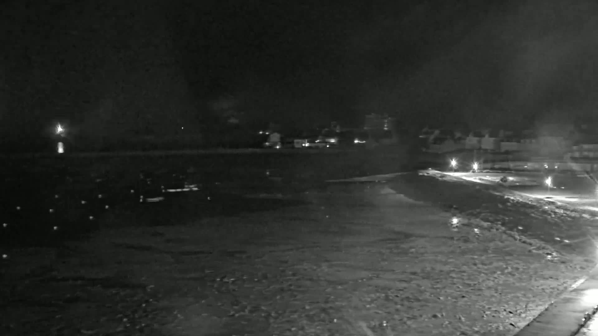 A nighttime, low-light view of a snow-covered waterfront area with dimly lit buildings across the water.