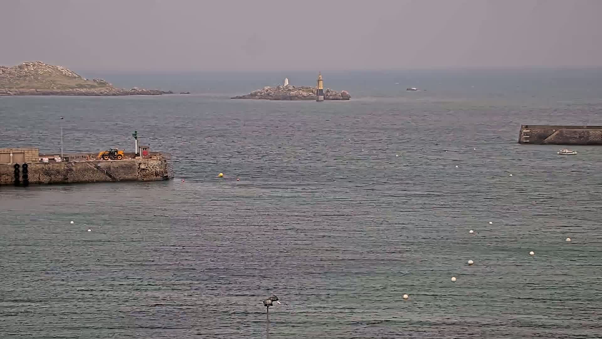 A harbor with a small yellow vehicle on a stone pier overlooks a calm, gray sea with a small island and lighthouse in the distance under an overcast sky.