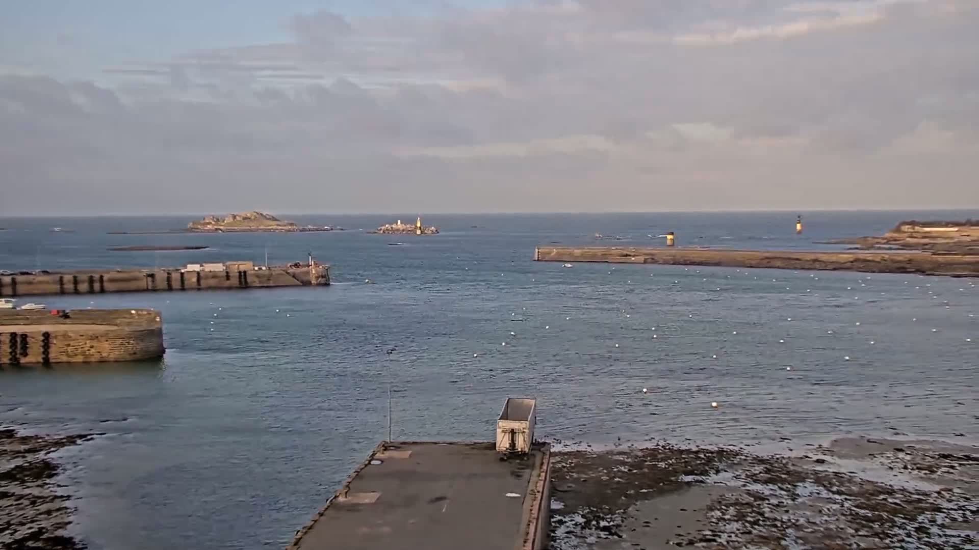 A harbor with stone walls and a docked trailer is viewed from above on a cloudy day, with islands and a lighthouse visible in the distance across calm water.