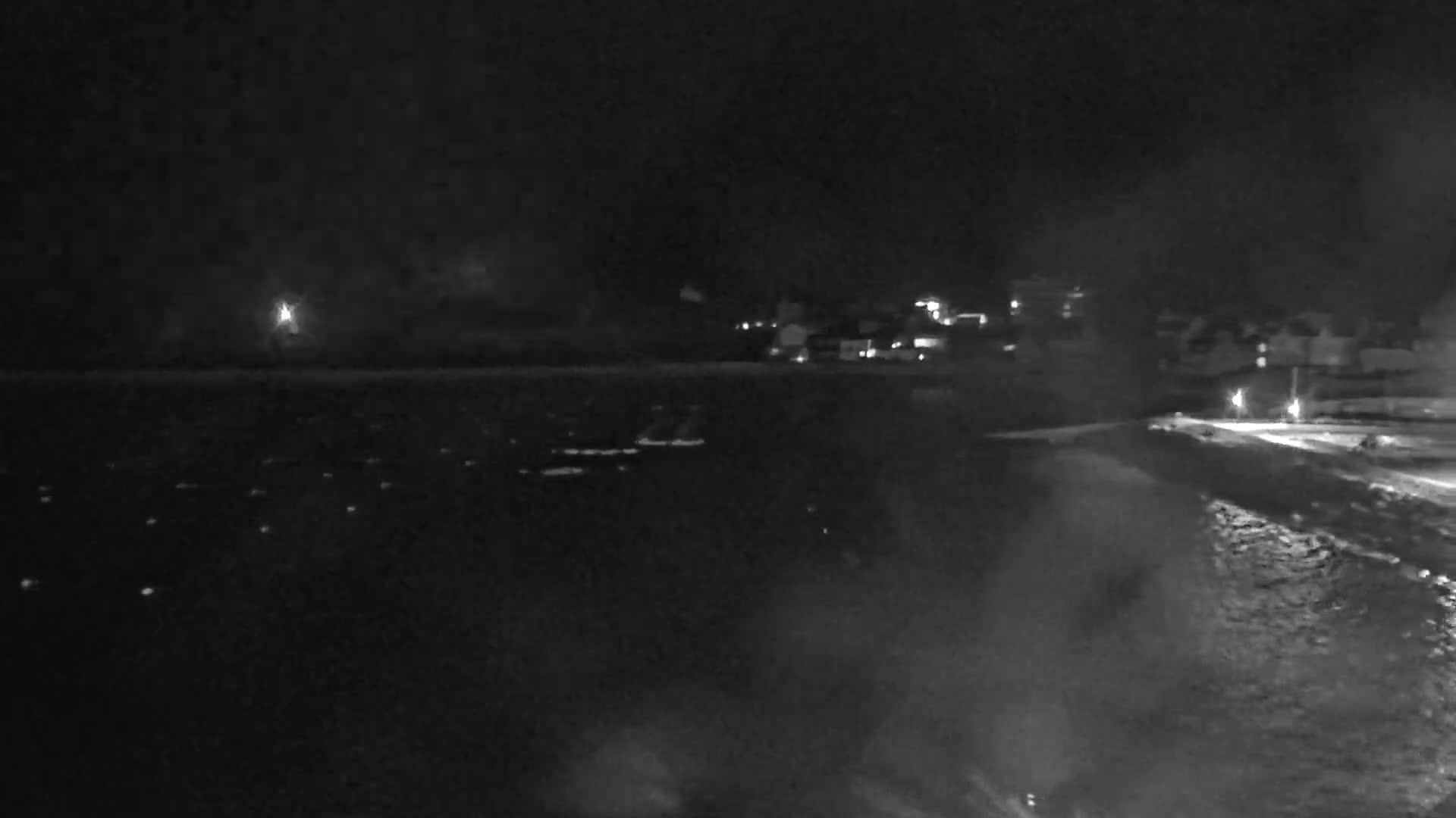 A nighttime, hazy view shows a dark body of water with faint lights scattered across its surface, a shoreline with dimly lit buildings, and a dark, overcast sky.