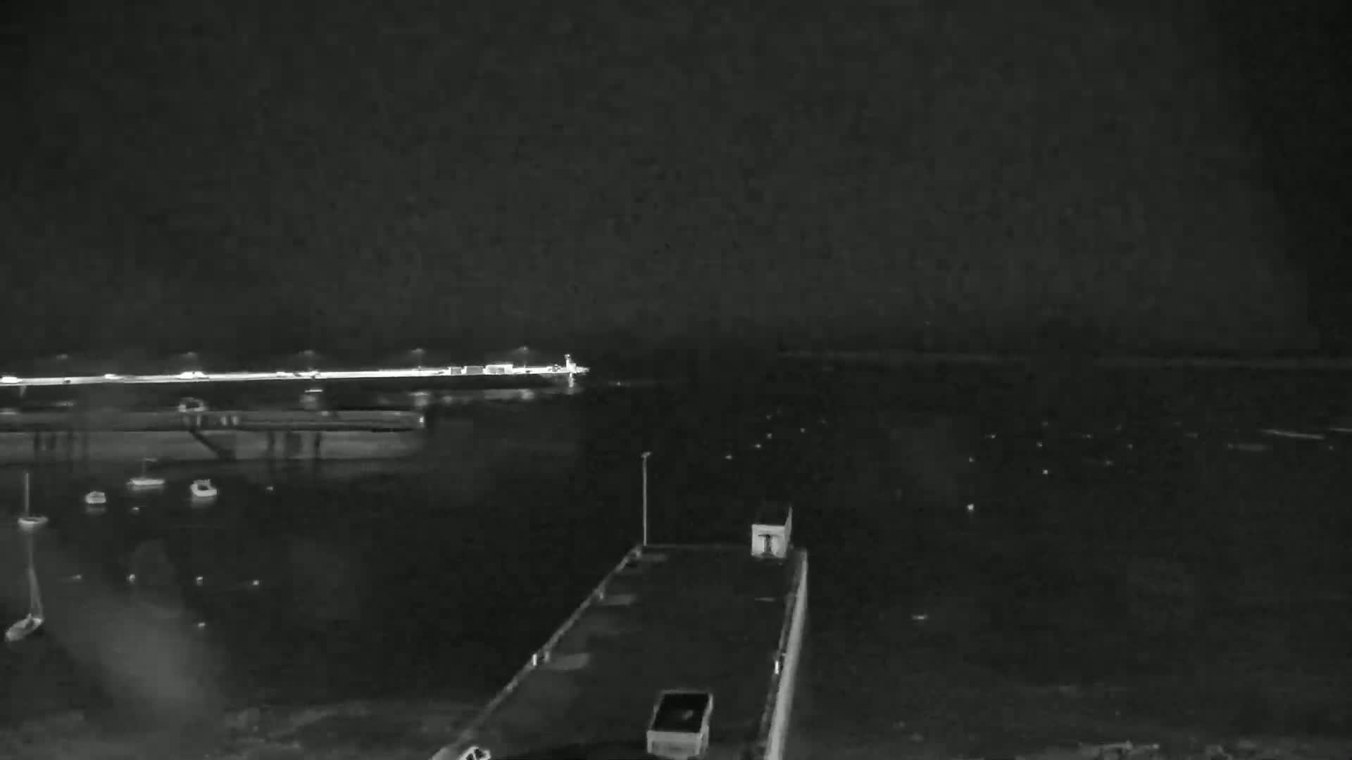 A nighttime view of a harbor with several boats, a pier with lights, and a dark body of water under a clear sky.