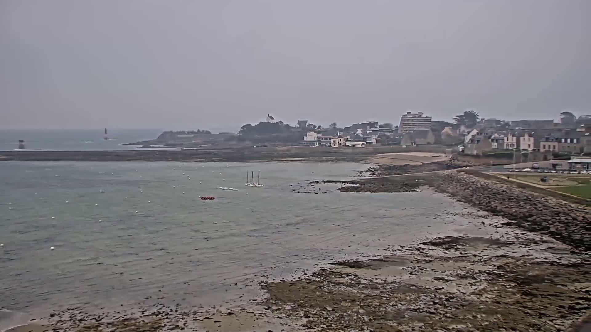 A hazy coastal scene shows a small town on the shore with a calm sea at low tide, and a small sailboat in the water.