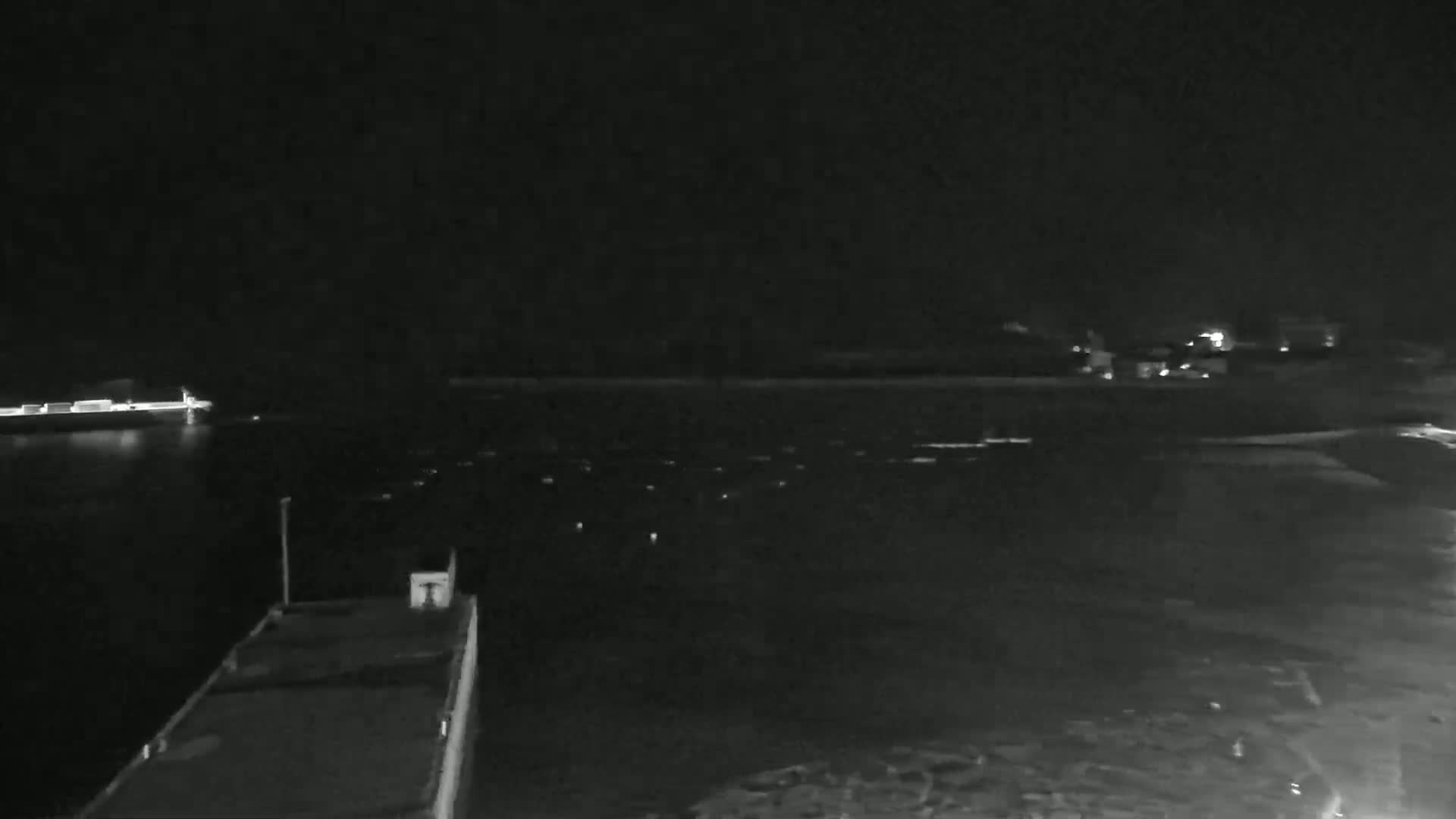 A dark nighttime scene shows a large barge or ship in the foreground on a calm body of water, with another vessel visible in the distance.