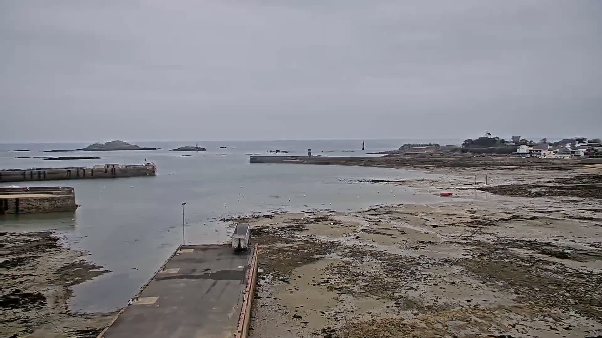 A harbor scene on an overcast day shows a small town on the horizon, a mostly exposed harbor bed, and a solitary truck parked near a paved area.