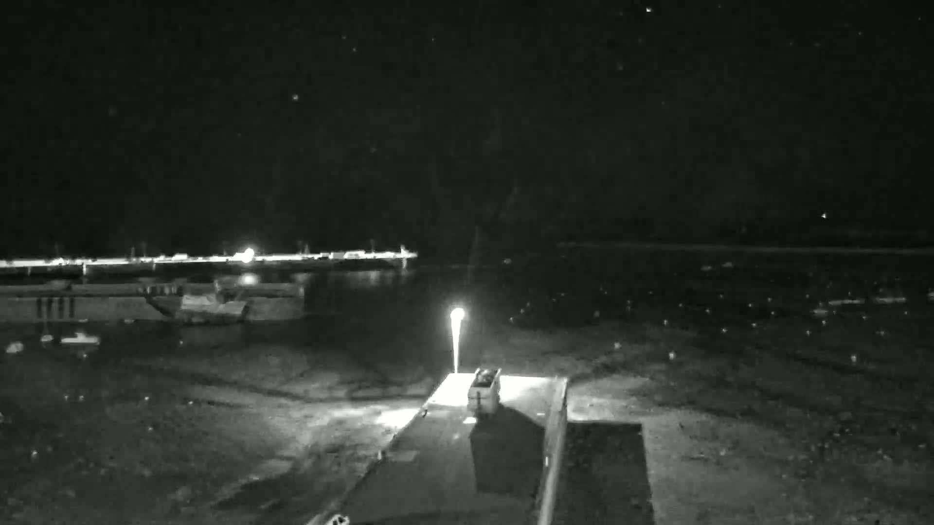 A nighttime, low-light view shows a vehicle parked near a lit lamppost in a mostly empty lot, with a body of water and distant lights visible in the background.