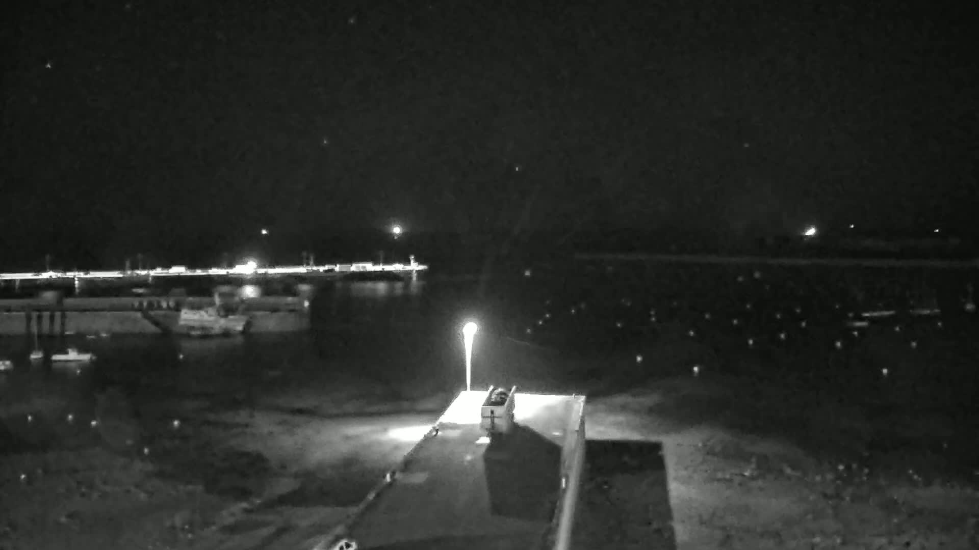A nighttime, low-light view of a harbor with a single light post illuminating a dark, flat area in the foreground, and a body of water with distant lights across it.