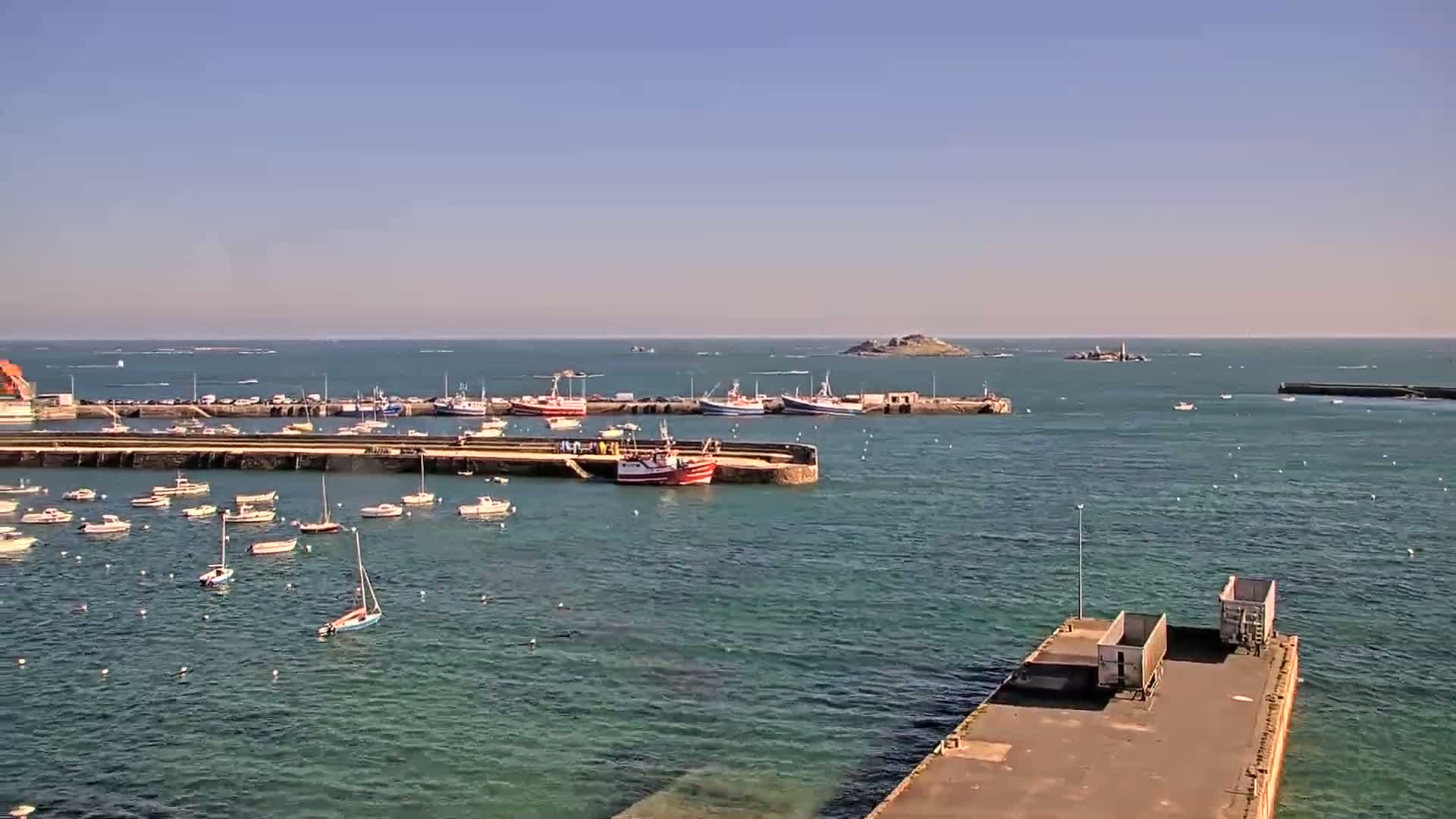 A harbor filled with numerous small boats and larger fishing vessels is seen on a sunny day, with a small island visible in the distance.