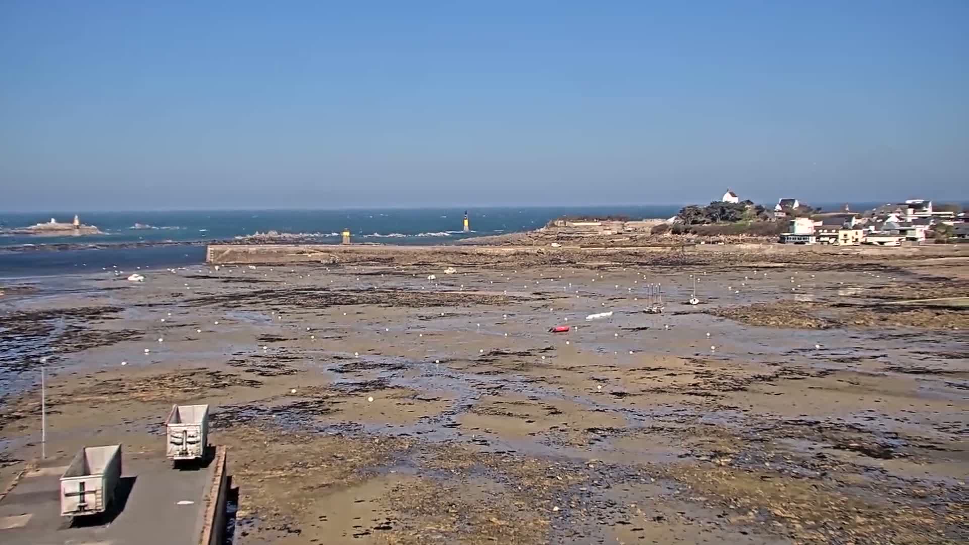 Under a clear blue sky, a wide expanse of mudflats reveals itself at low tide, dotted with small boats and buoys, near a coastline featuring a small village and distant lighthouses.