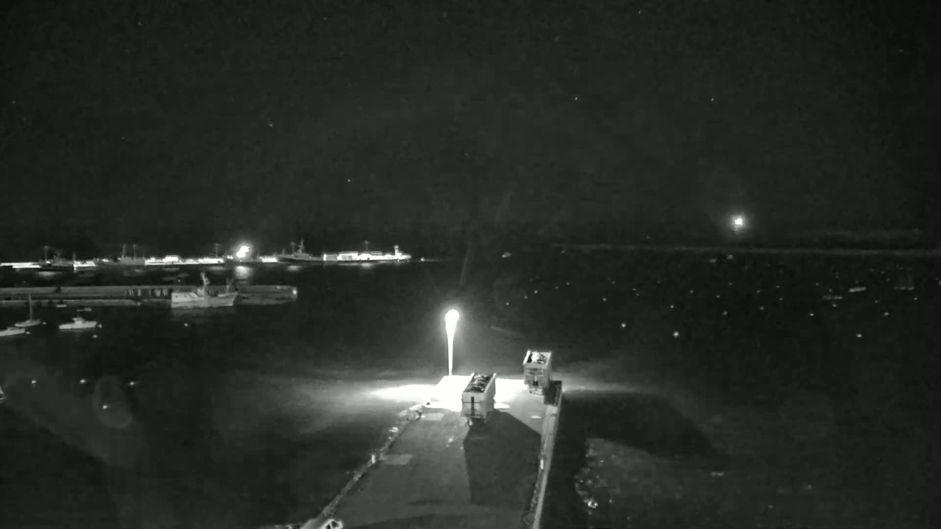 A nighttime, high-angle view shows a harbor with several boats, a pier with two vehicles, and a bright light illuminating the scene.