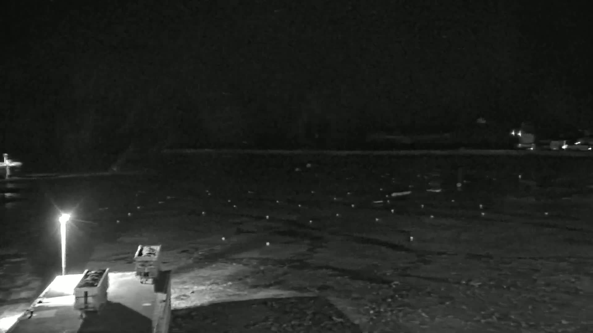 A nighttime, low-light view shows a dark, expansive area with numerous small, spaced lights, a single lamppost, and a dark structure in the bottom left corner.