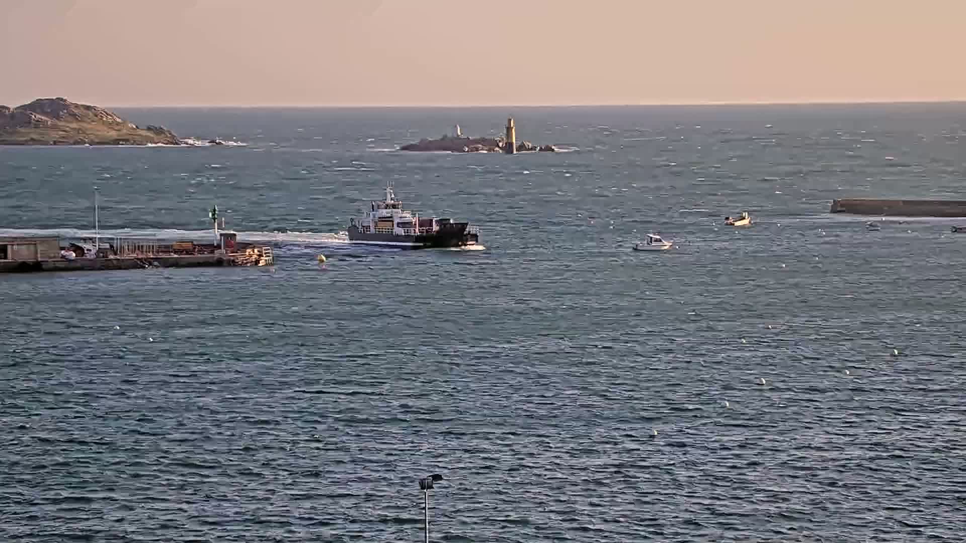 A ferry passes by a small harbor on a windy day, with other small boats and a distant island with a lighthouse visible in the choppy water.