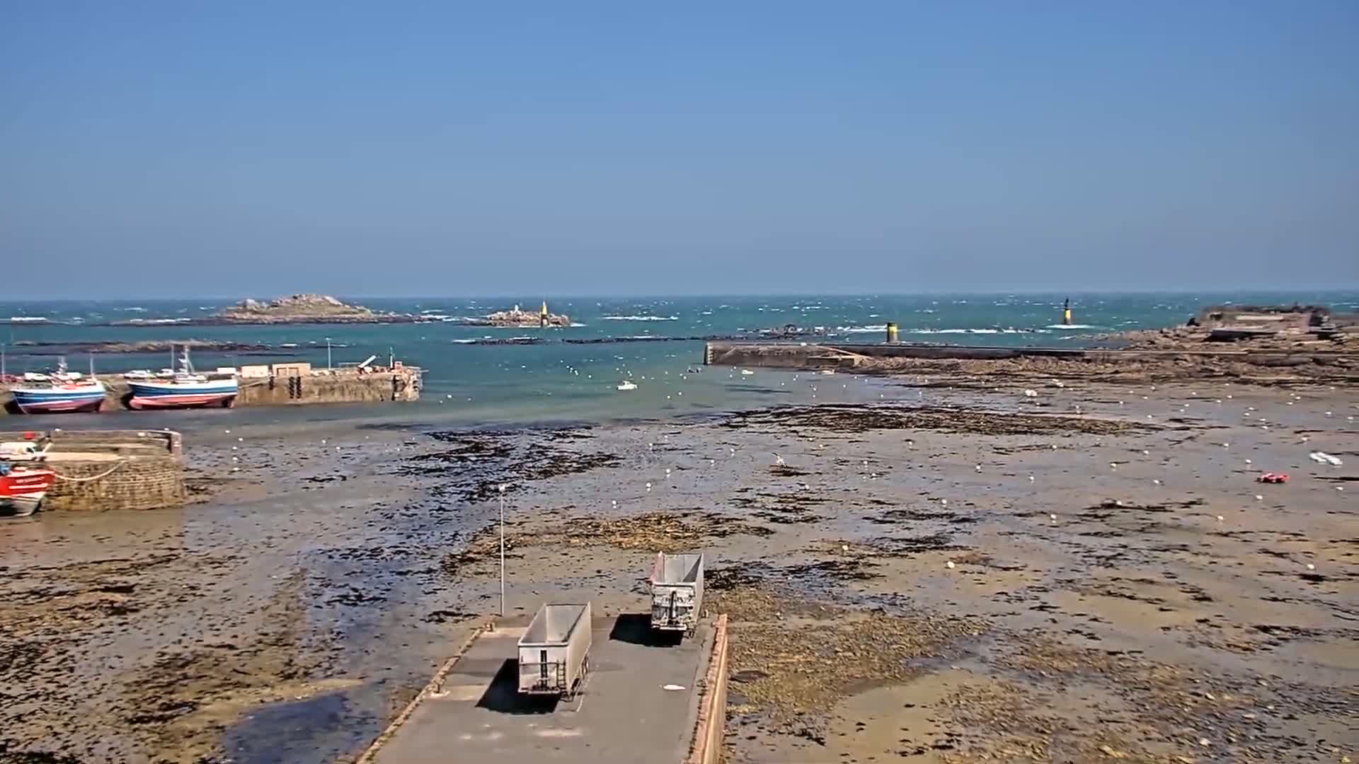 A harbor with several boats docked, a rocky shoreline exposed at low tide under a sunny, windy sky, and two trailers parked on a paved area near the water.