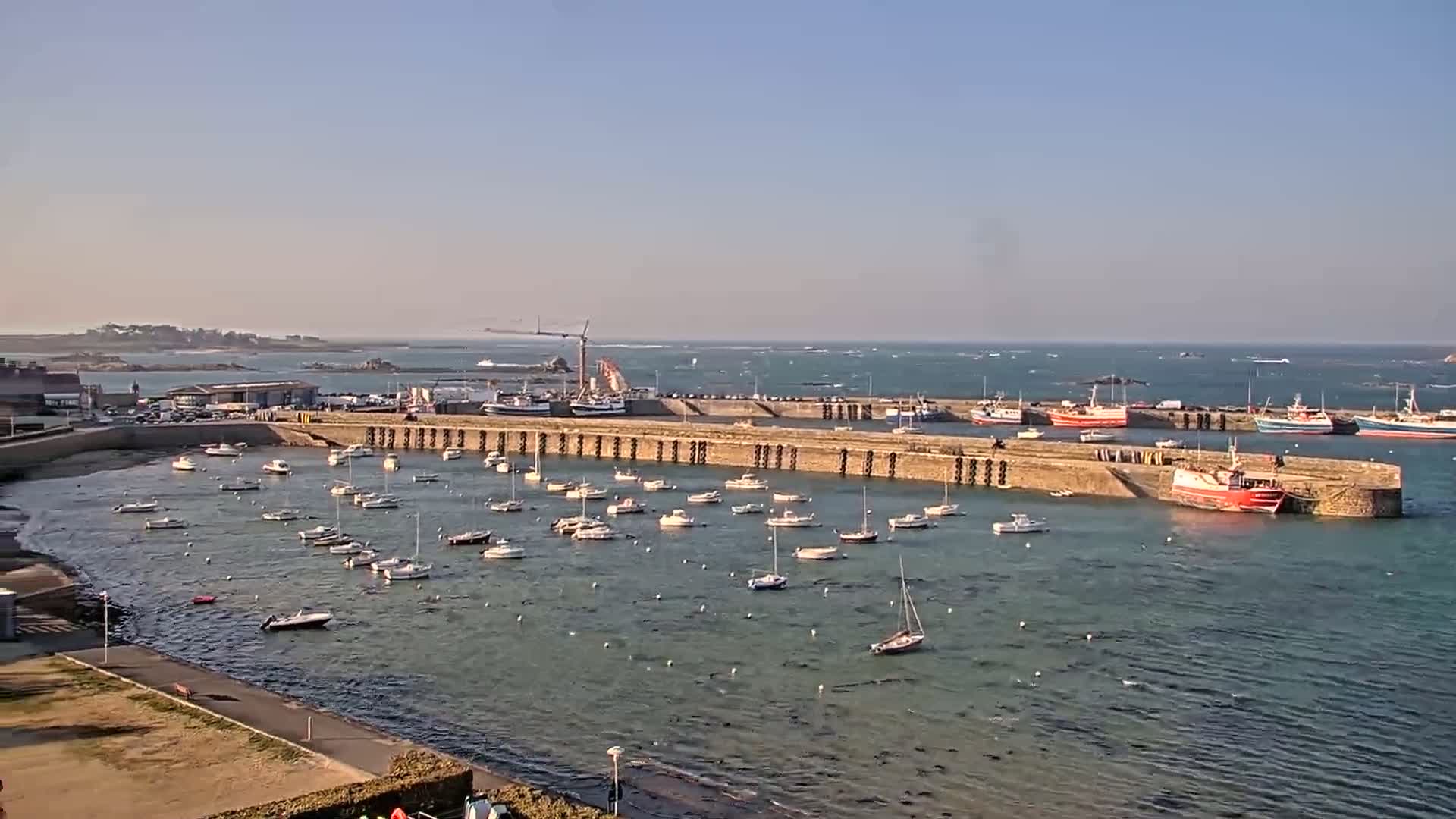 A calm harbor filled with numerous small boats and larger fishing vessels is enclosed by a stone wall under a clear, sunny sky.