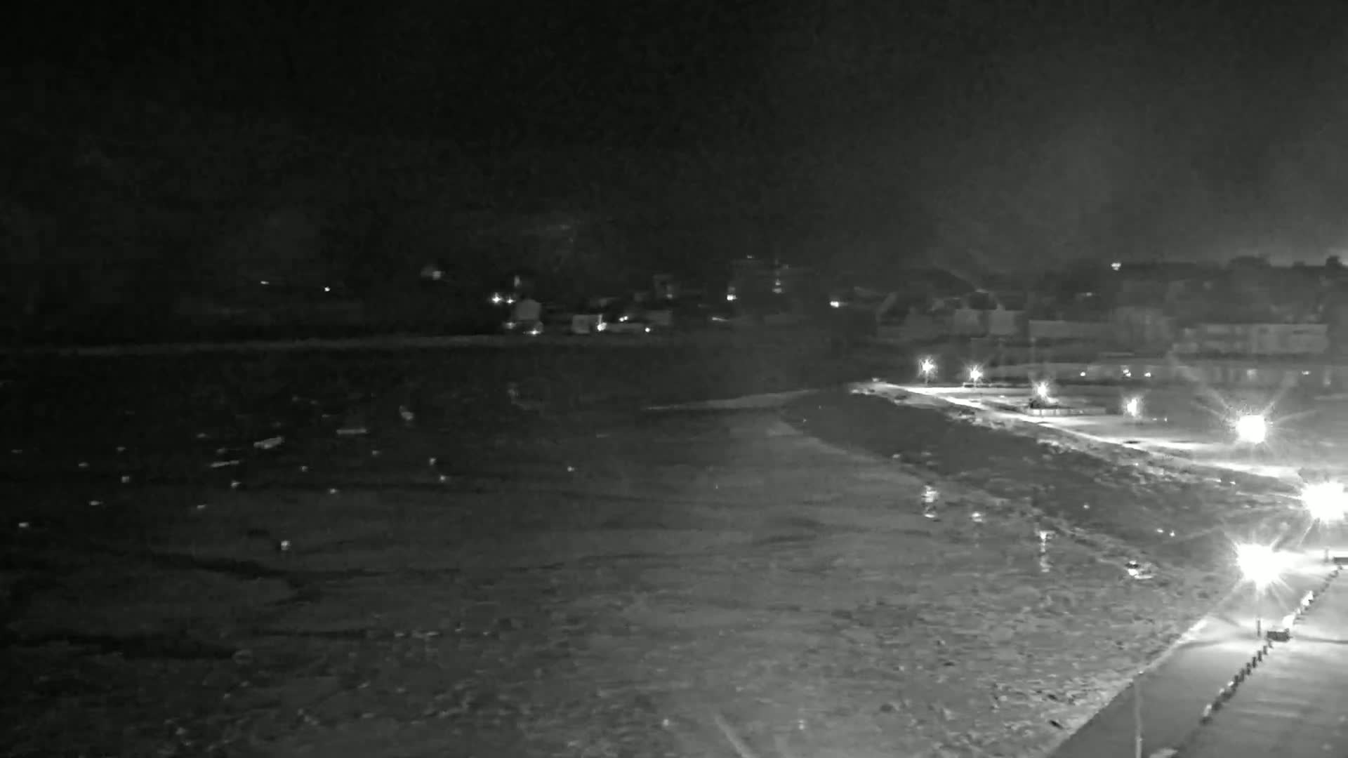A nighttime, low-light view of a dark, rough body of water lapping at a shoreline with lights illuminating a paved walkway and buildings in the distance.