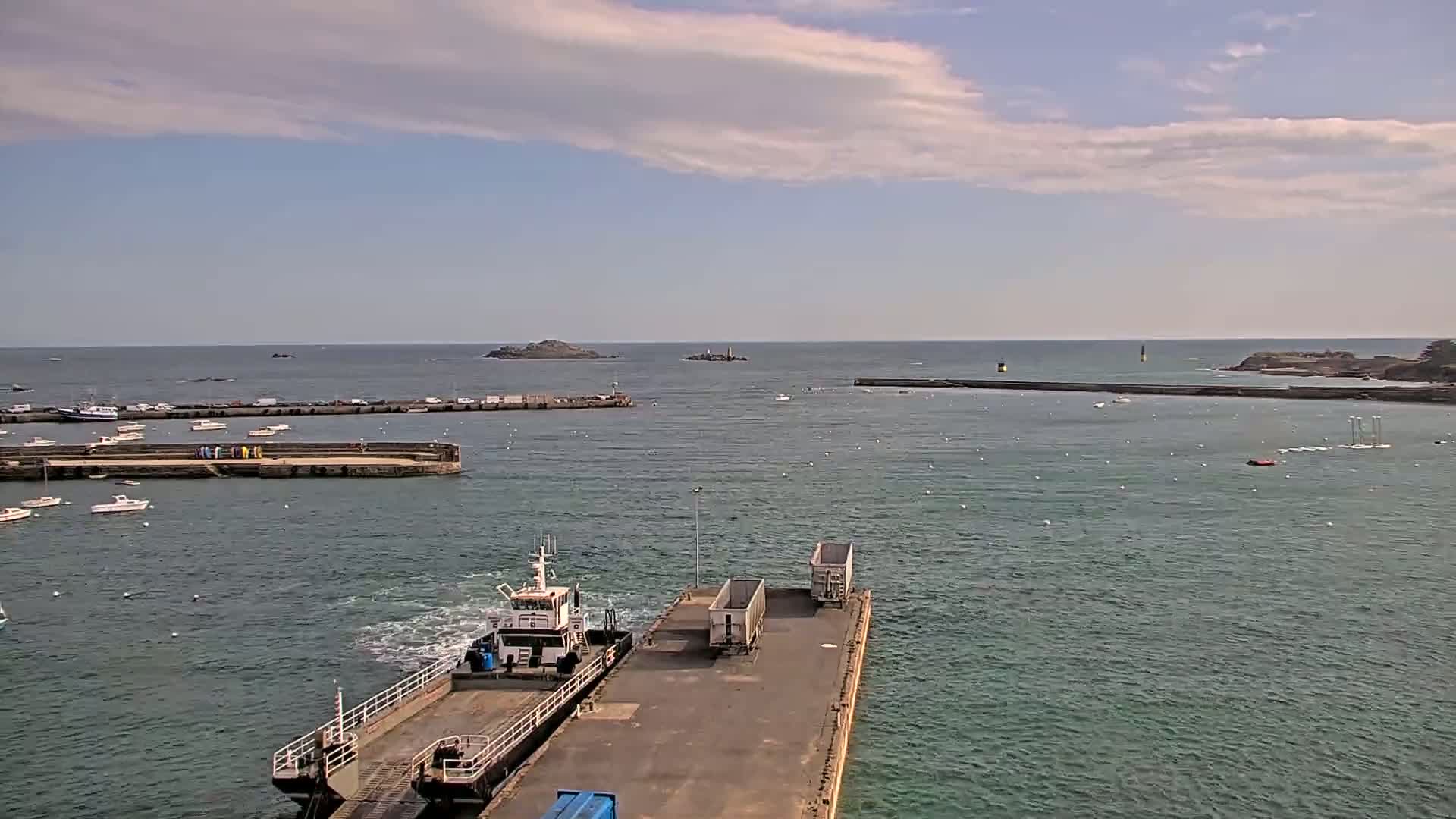 A tugboat approaches a dock with two shipping containers under partly cloudy skies, with calm waters and several small boats in the harbor.