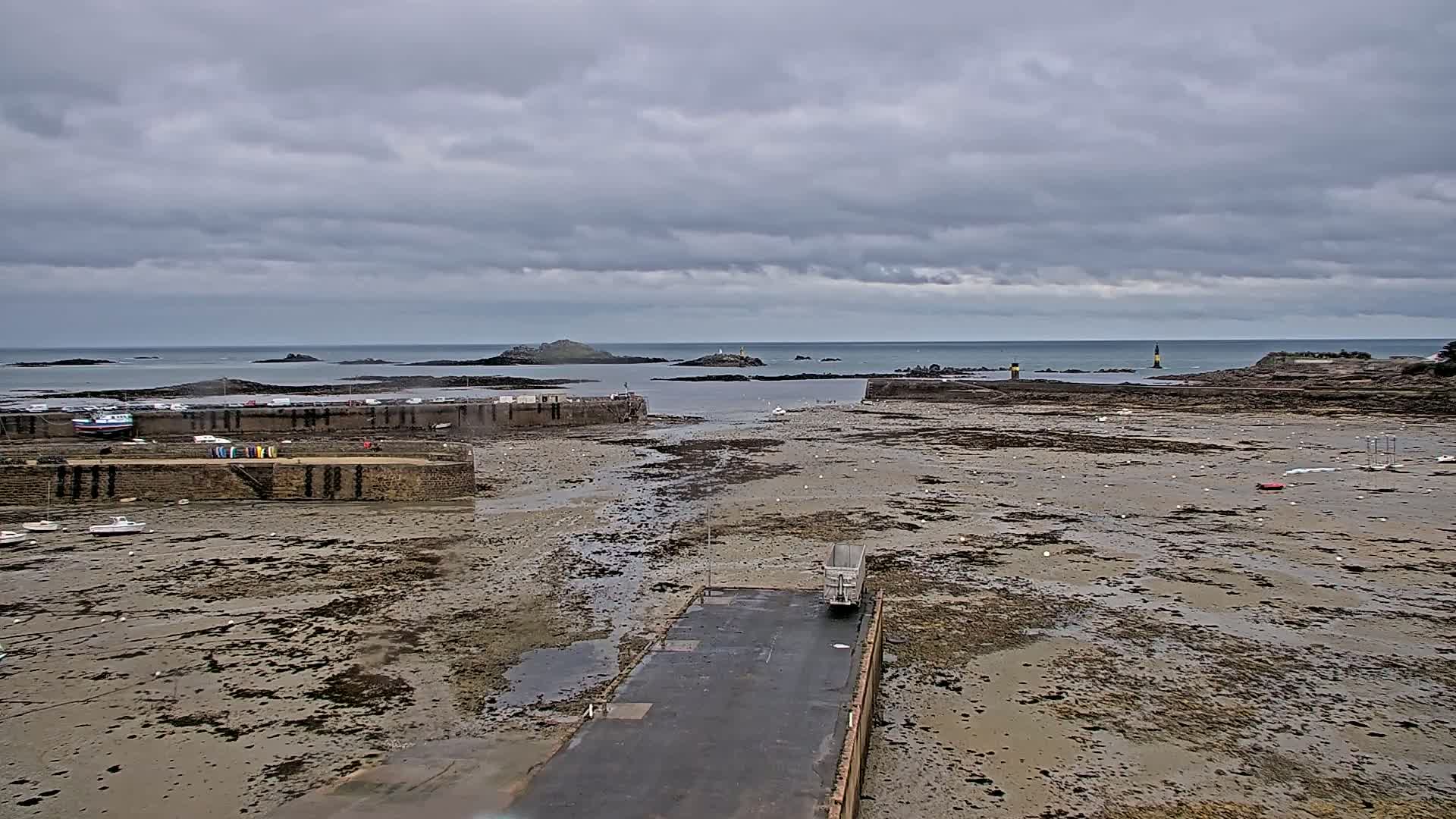A mostly exposed harbor with a few boats, a truck, and distant islands under a cloudy sky.