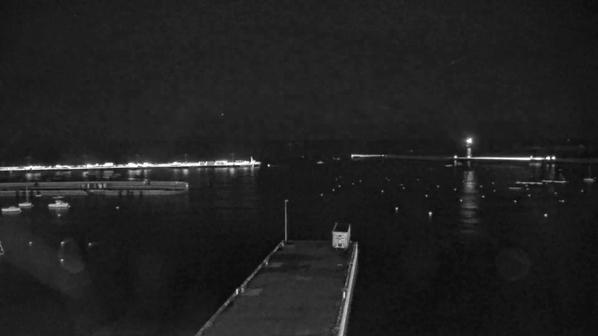 A nighttime view of a harbor with boats moored, a lit-up pier, and a lighthouse in the distance, under a dark, clear sky.
