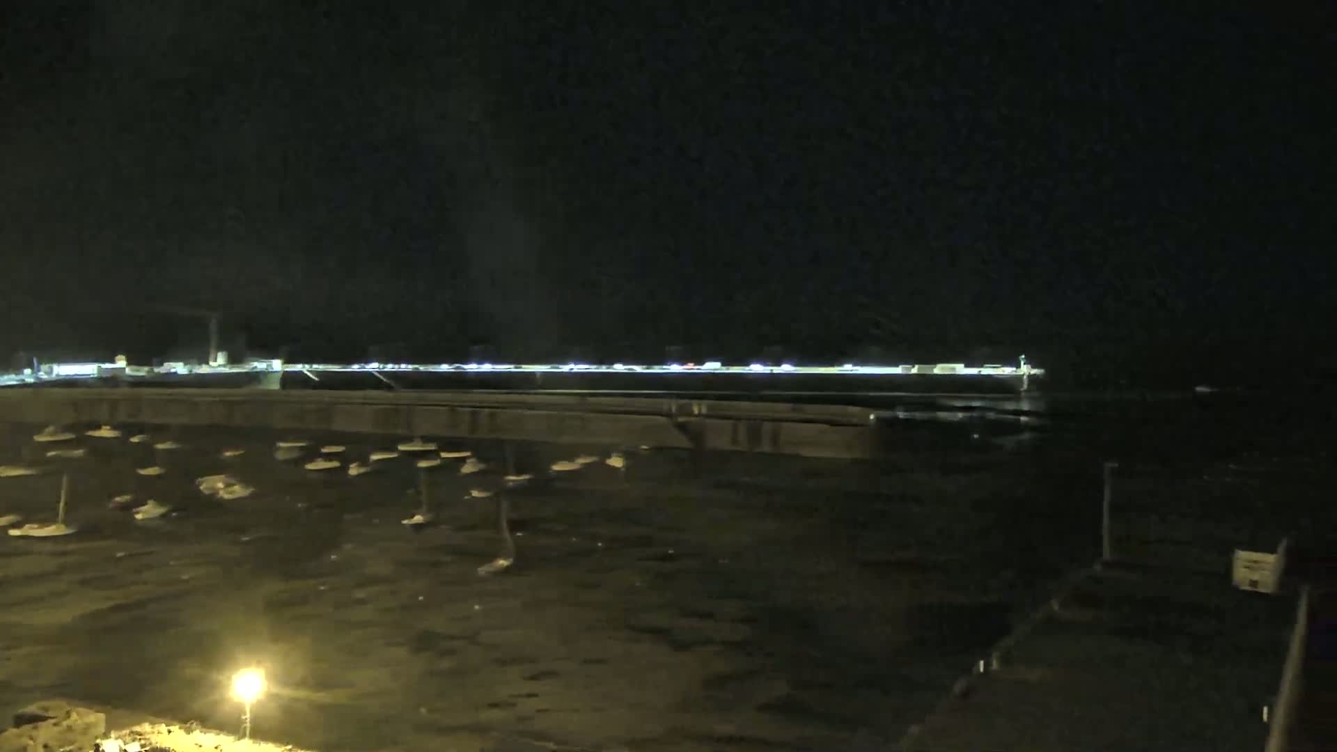 A nighttime view shows a harbor filled with numerous small boats, a distant shoreline with lights, and a dark sky.