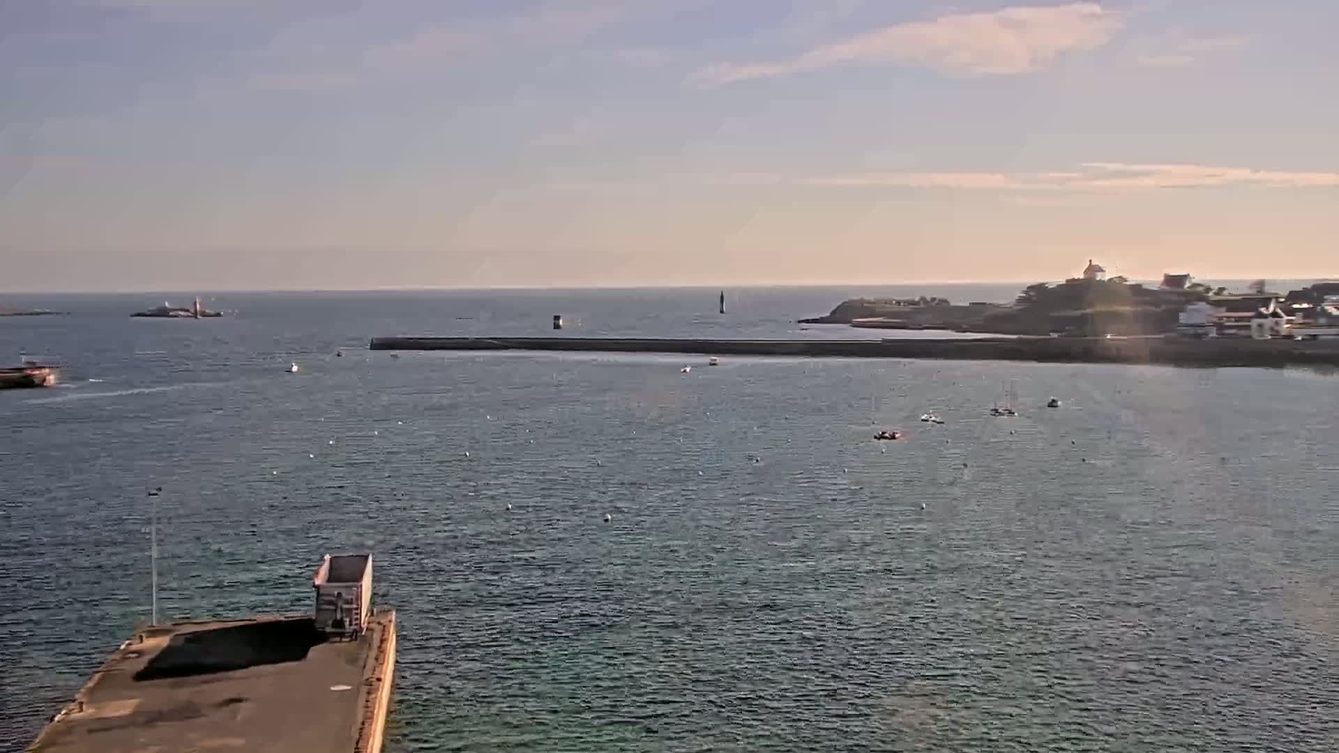 A calm body of water with several small boats and a distant landmass under a partly cloudy sky shows a truck parked on a pier in the foreground.
