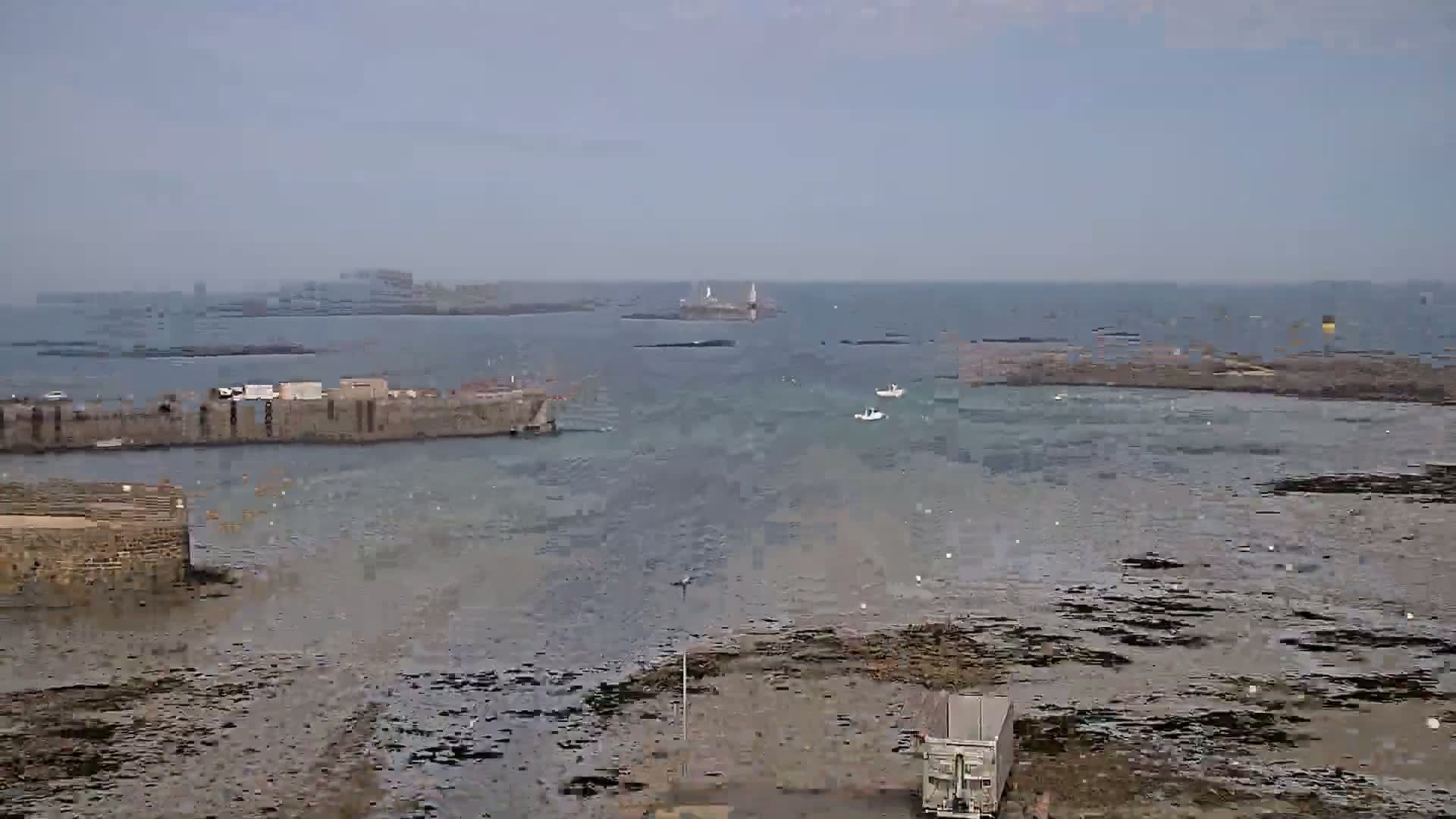 A harbor scene on a calm, overcast day shows a stone seawall, a few small boats, and exposed rocks at low tide, with distant islands visible on the horizon.