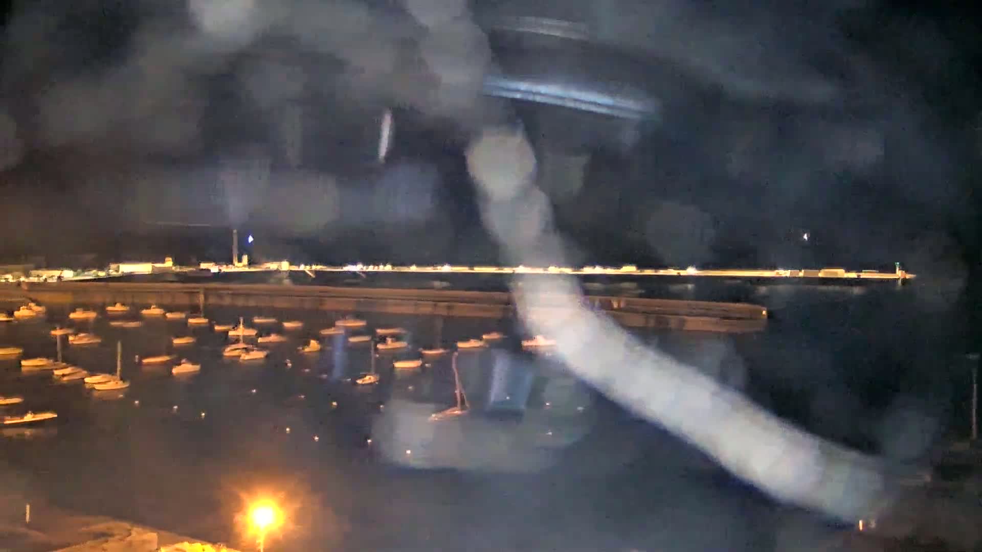 A nighttime view of a harbor filled with numerous boats, seen through a rain-streaked window or similar obscuring surface.