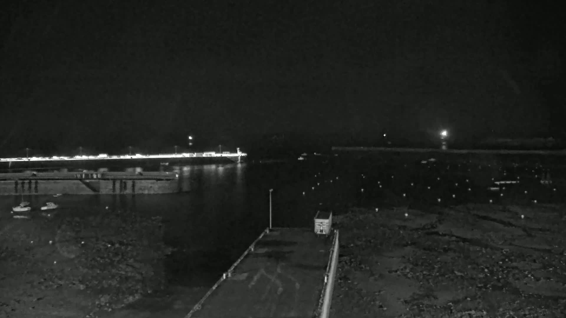A nighttime view of a harbor with several boats, a pier, and a distant shoreline illuminated by lights.