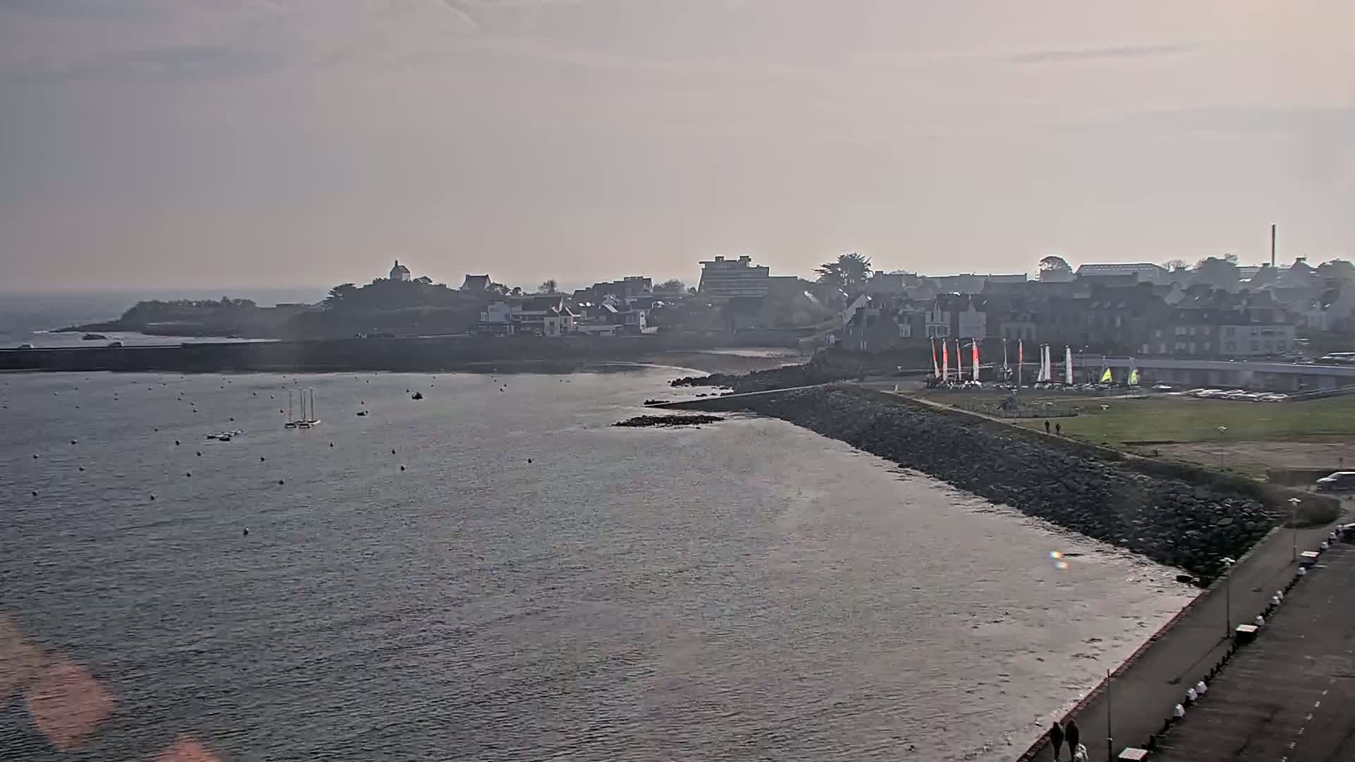 A hazy, overcast day reveals a coastal town with a calm bay, several sailboats, and a rocky seawall.