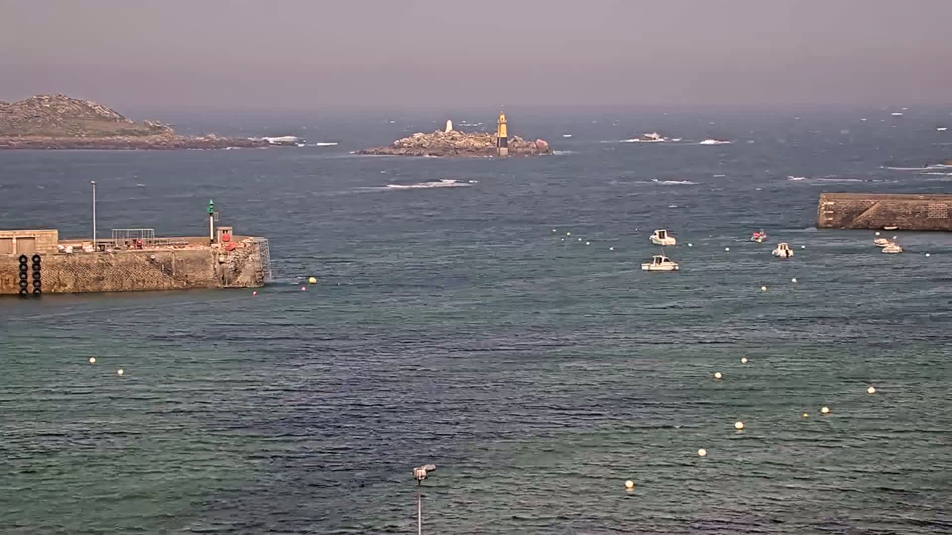 A rocky island with a lighthouse is visible in the distance across a choppy sea, where several small boats are anchored near a harbor wall.