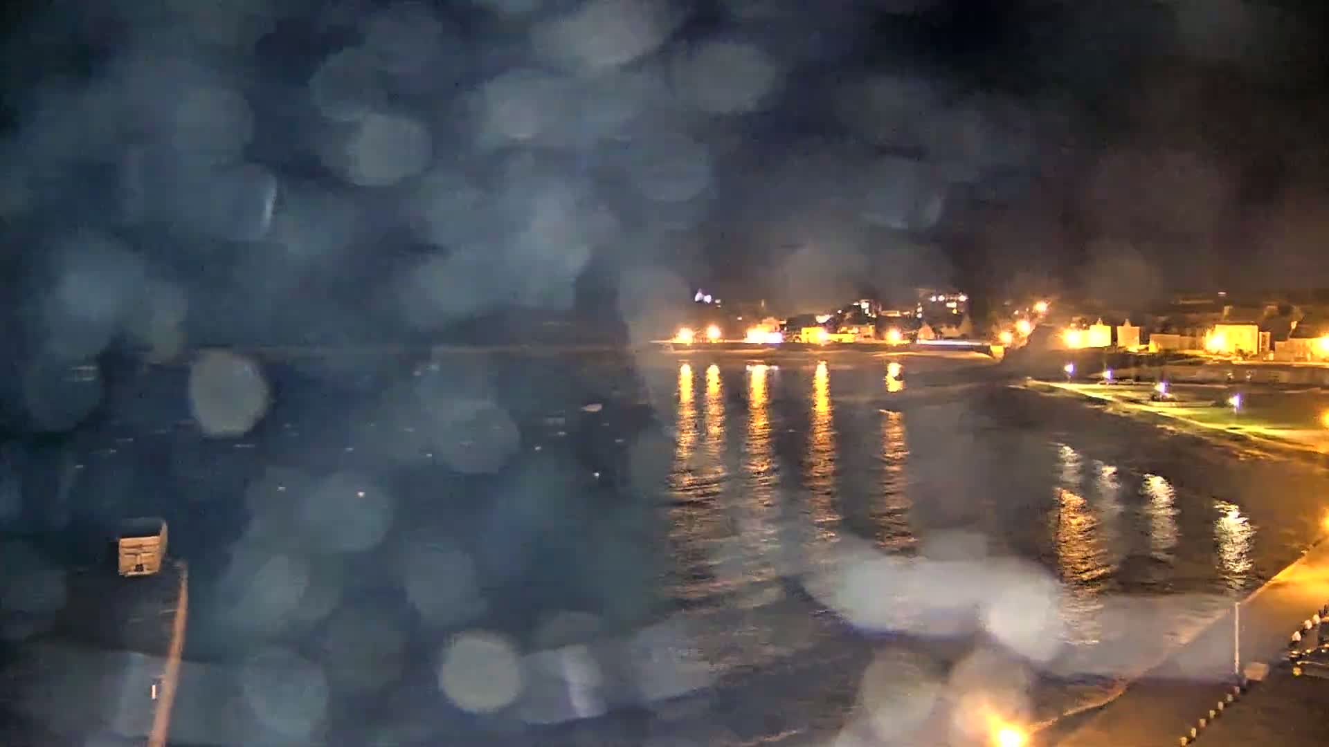 A nighttime view of a calm, dark ocean reflecting the lights of a coastal town under a rain-blurred sky.