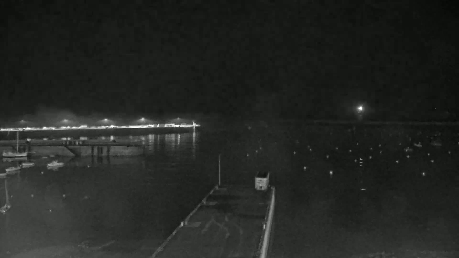 A nighttime view of a harbor with boats moored at the docks, under a dark sky with a few faint lights visible in the distance.