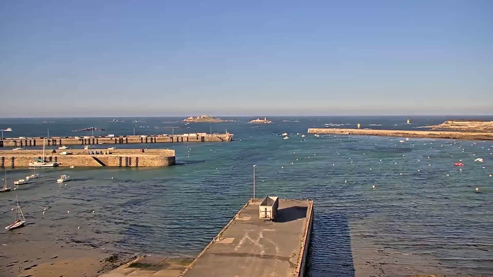 A harbor with numerous boats, stone walls, and a distant island is visible on a clear, sunny day.