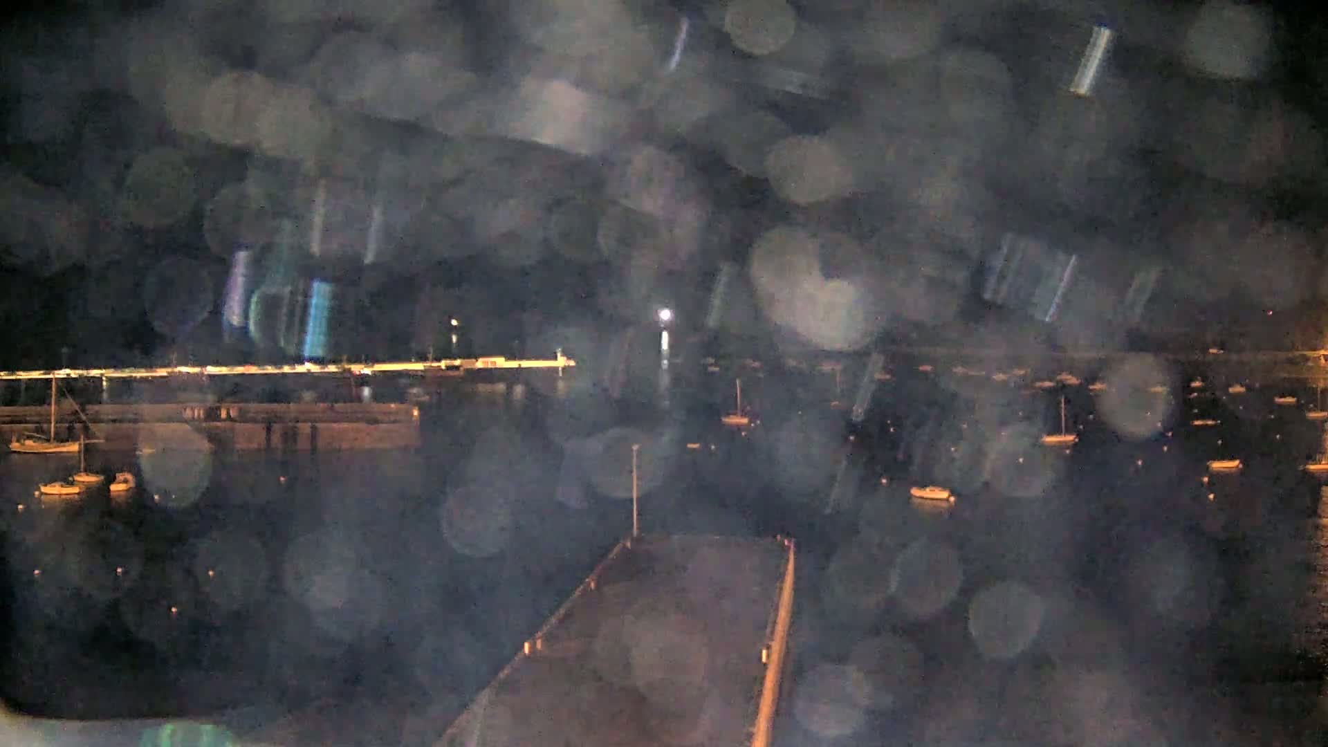 A nighttime view of a harbor filled with numerous boats, seen through a rain-streaked window or lens.