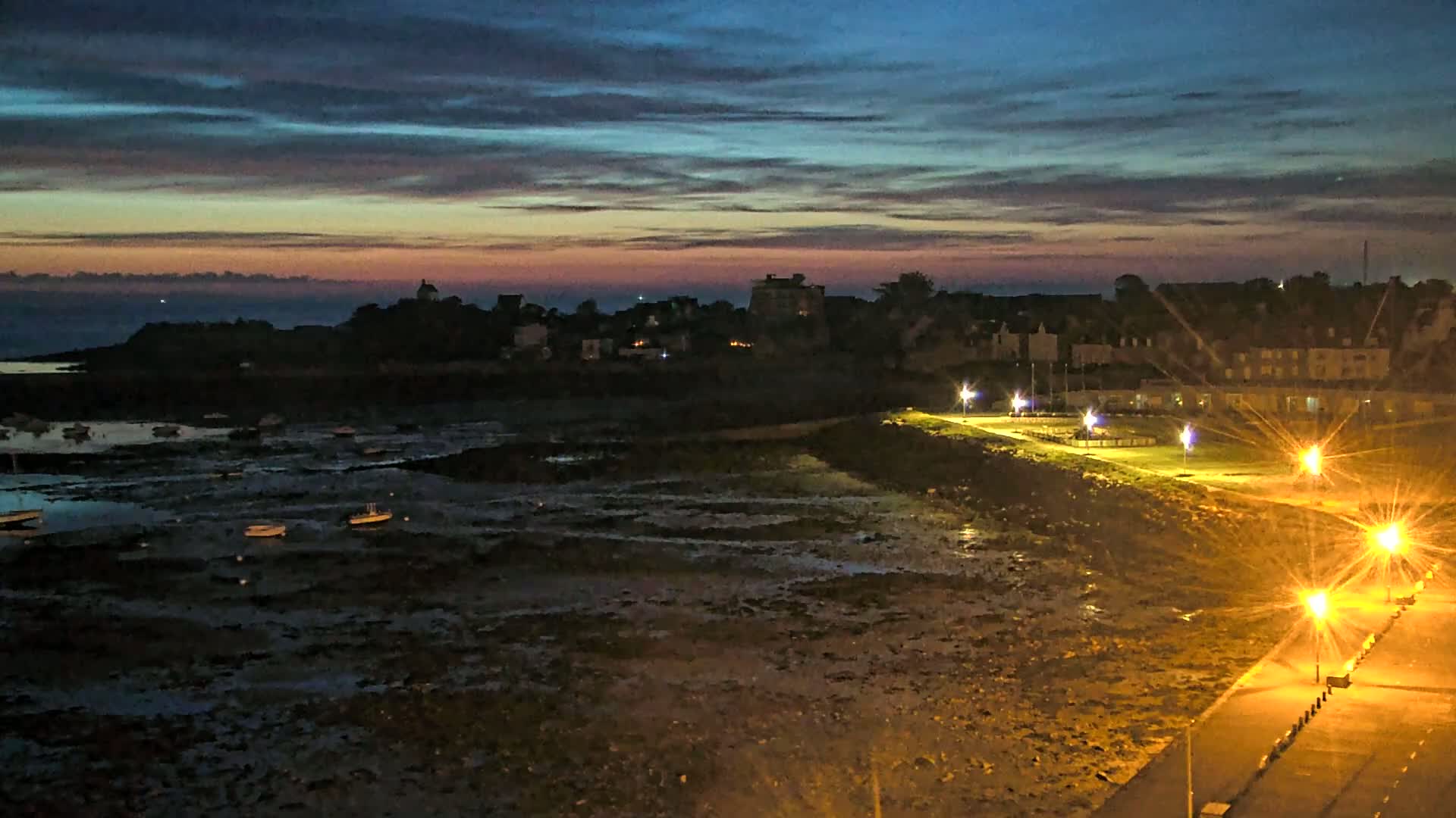 A coastal town at night, under a twilight sky with colorful clouds, shows a low-tide harbor with several small boats and illuminated waterfront pathways.