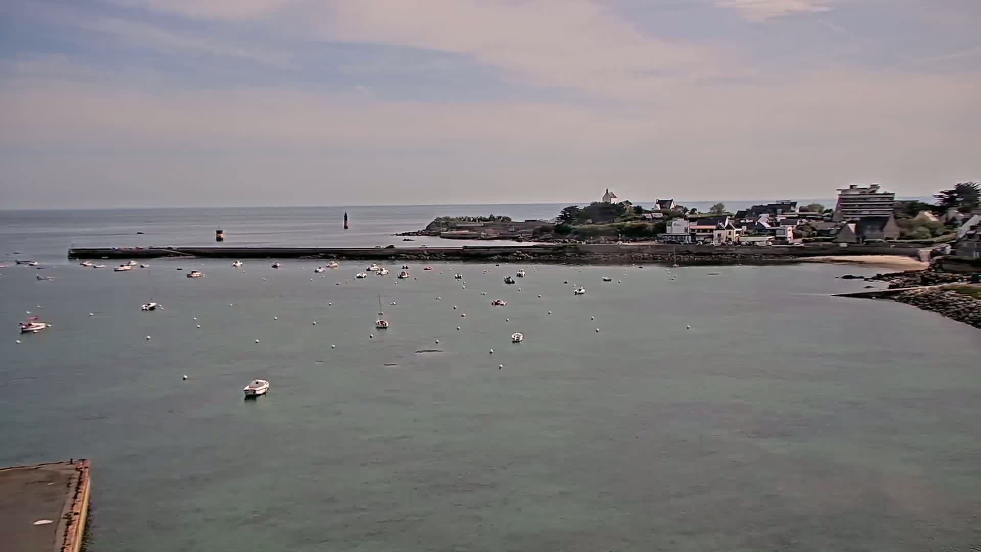 A calm bay with numerous small boats is nestled next to a small town under a mostly cloudy sky.