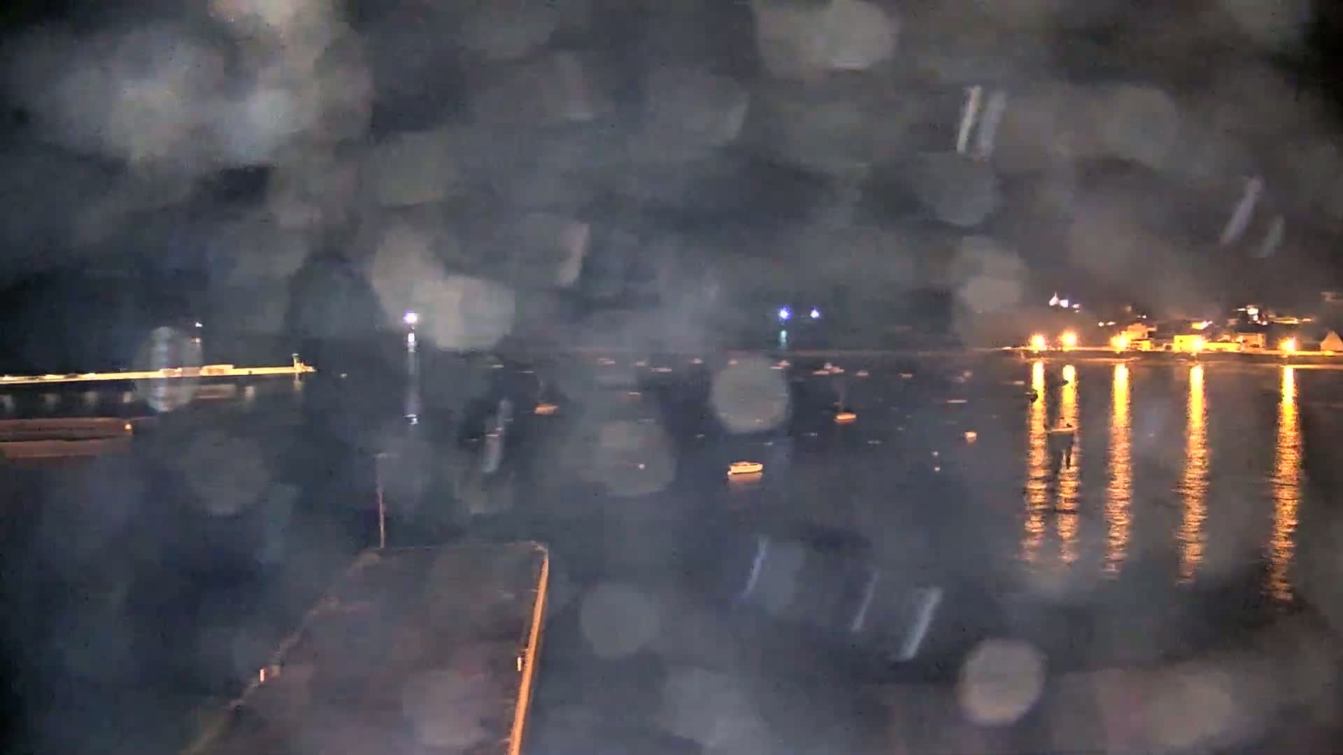 A nighttime view of a harbor with numerous boats and a town's lights reflecting on the water, obscured by rain or mist.