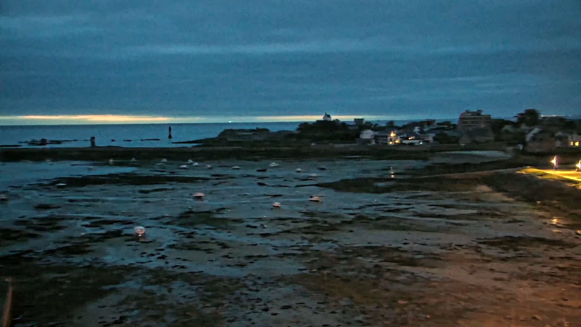 Under a dark, cloudy sky, a coastal town is visible at low tide, with numerous small boats resting on the exposed seabed.