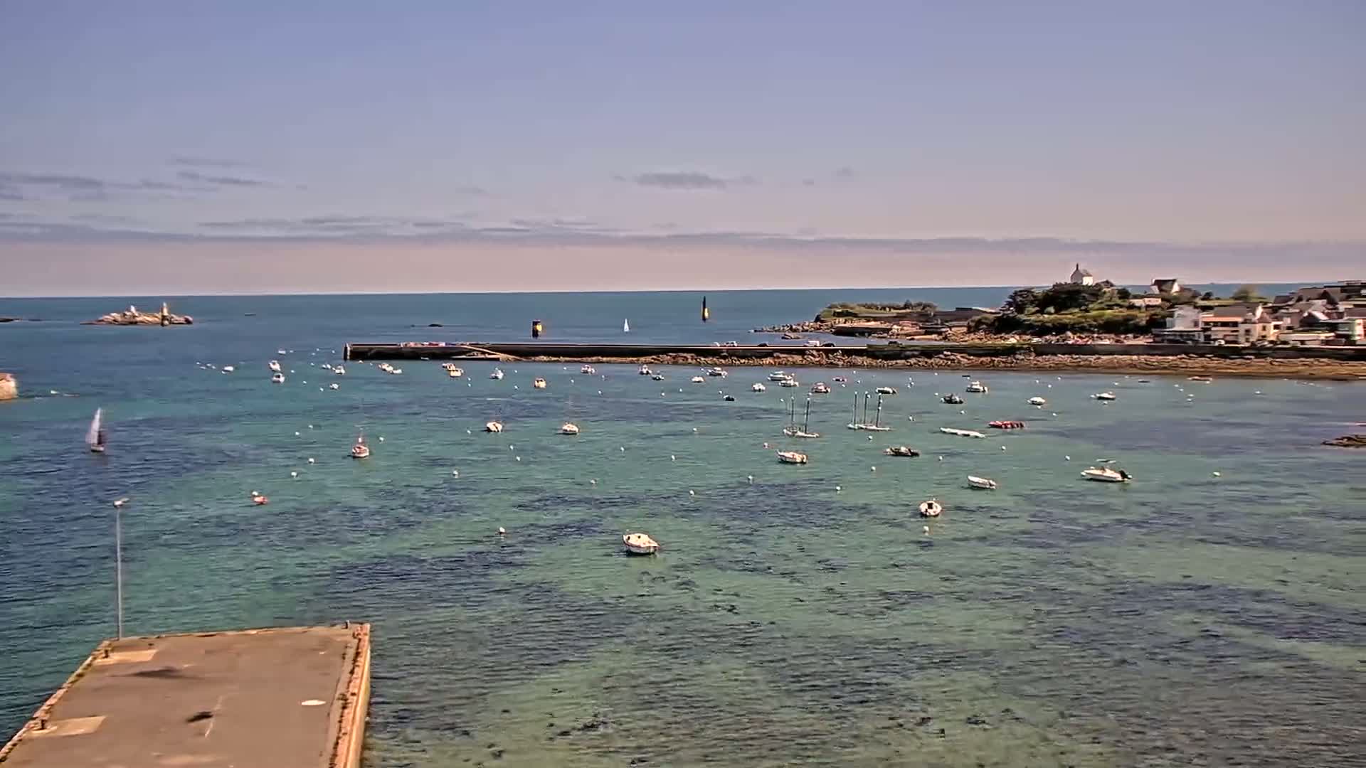 A calm, clear day reveals a harbor filled with numerous small boats, anchored near a seaside town nestled beside a rocky coastline.