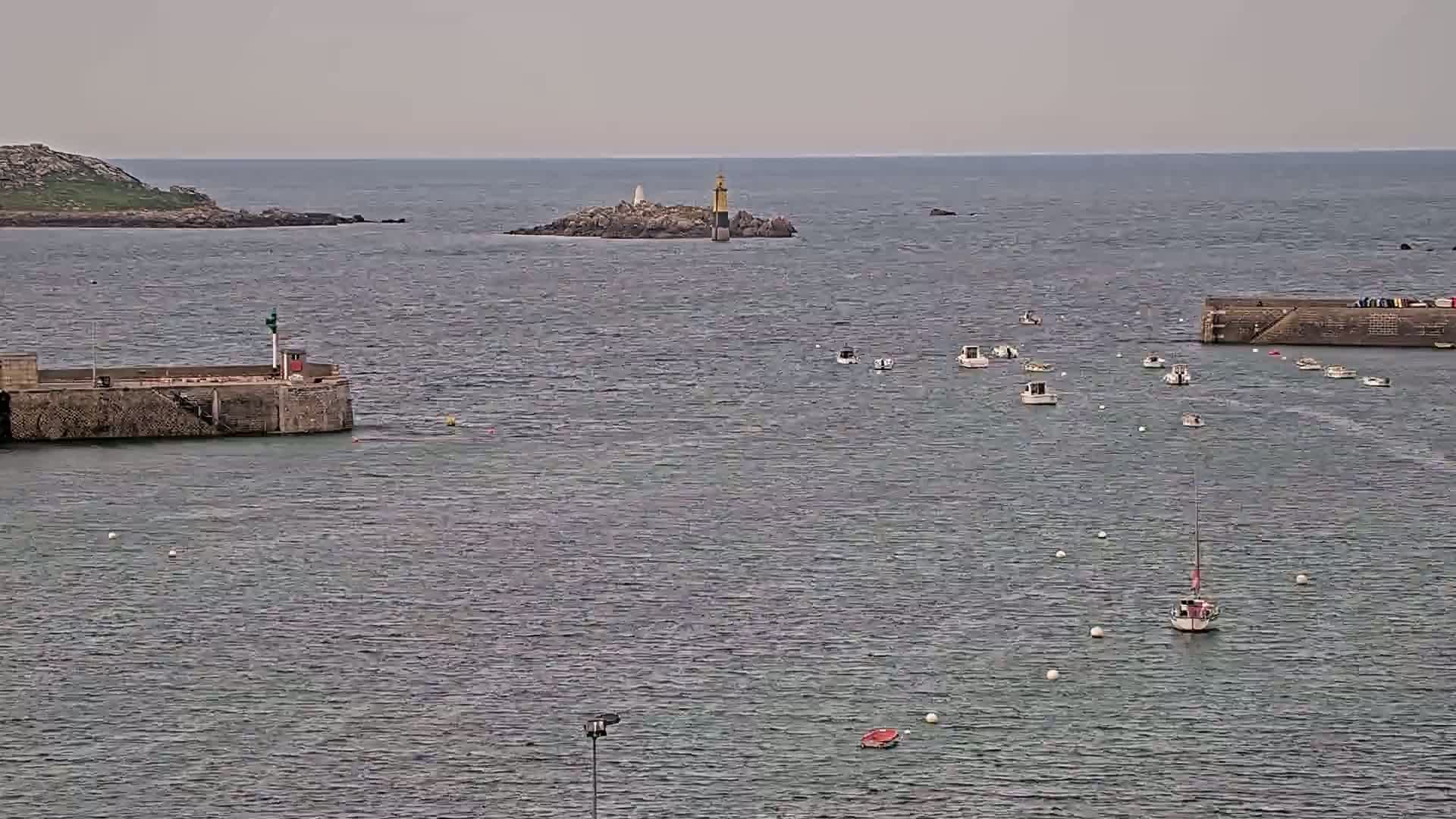 A calm, gray day shows numerous small boats in a harbor near a small island with a lighthouse, and a larger stone structure is visible on the left.