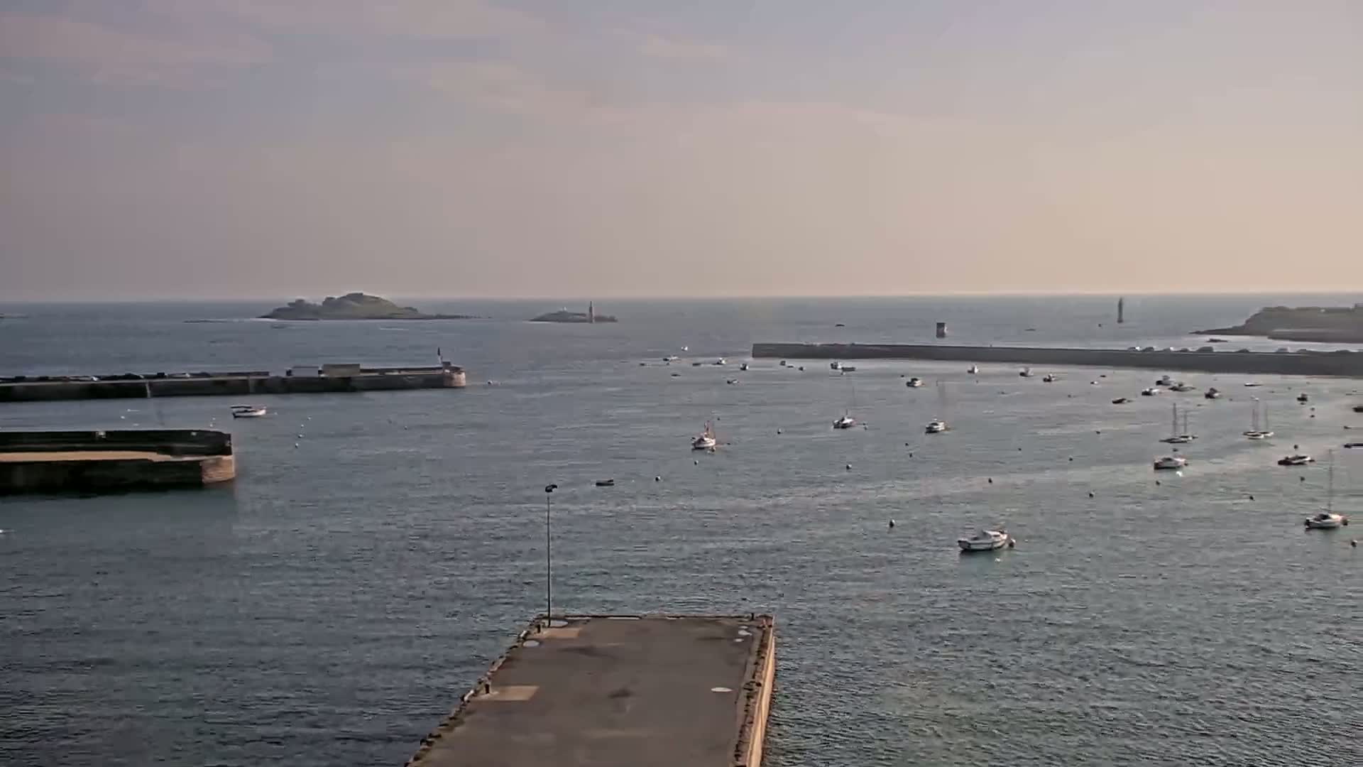 A calm harbor with numerous small boats is seen from an elevated vantage point on a partly sunny day, with islands and a lighthouse visible in the distance.