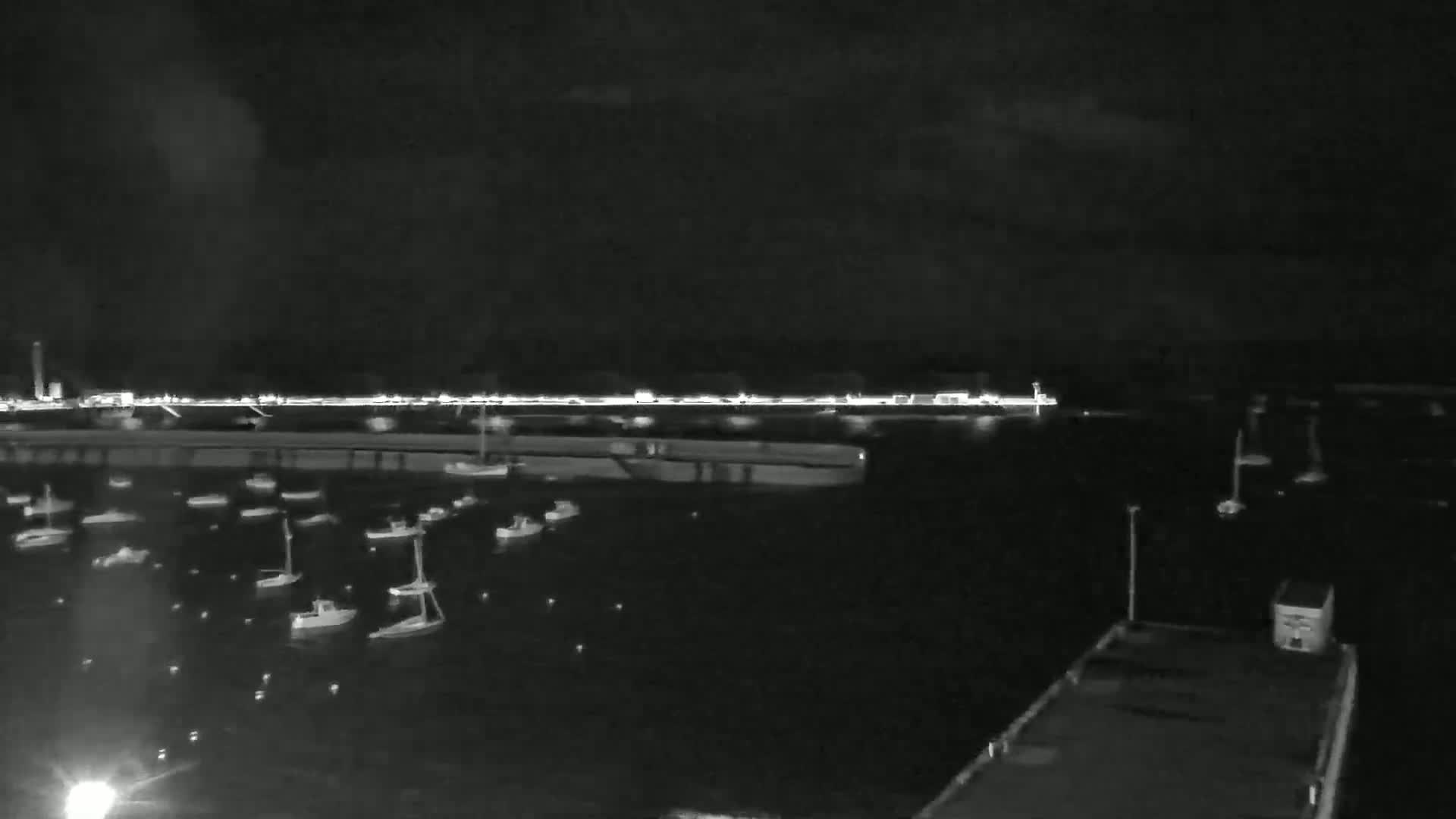 At night, numerous small boats are moored in a dark harbor, with a brightly lit waterfront visible in the distance under a cloudy sky.