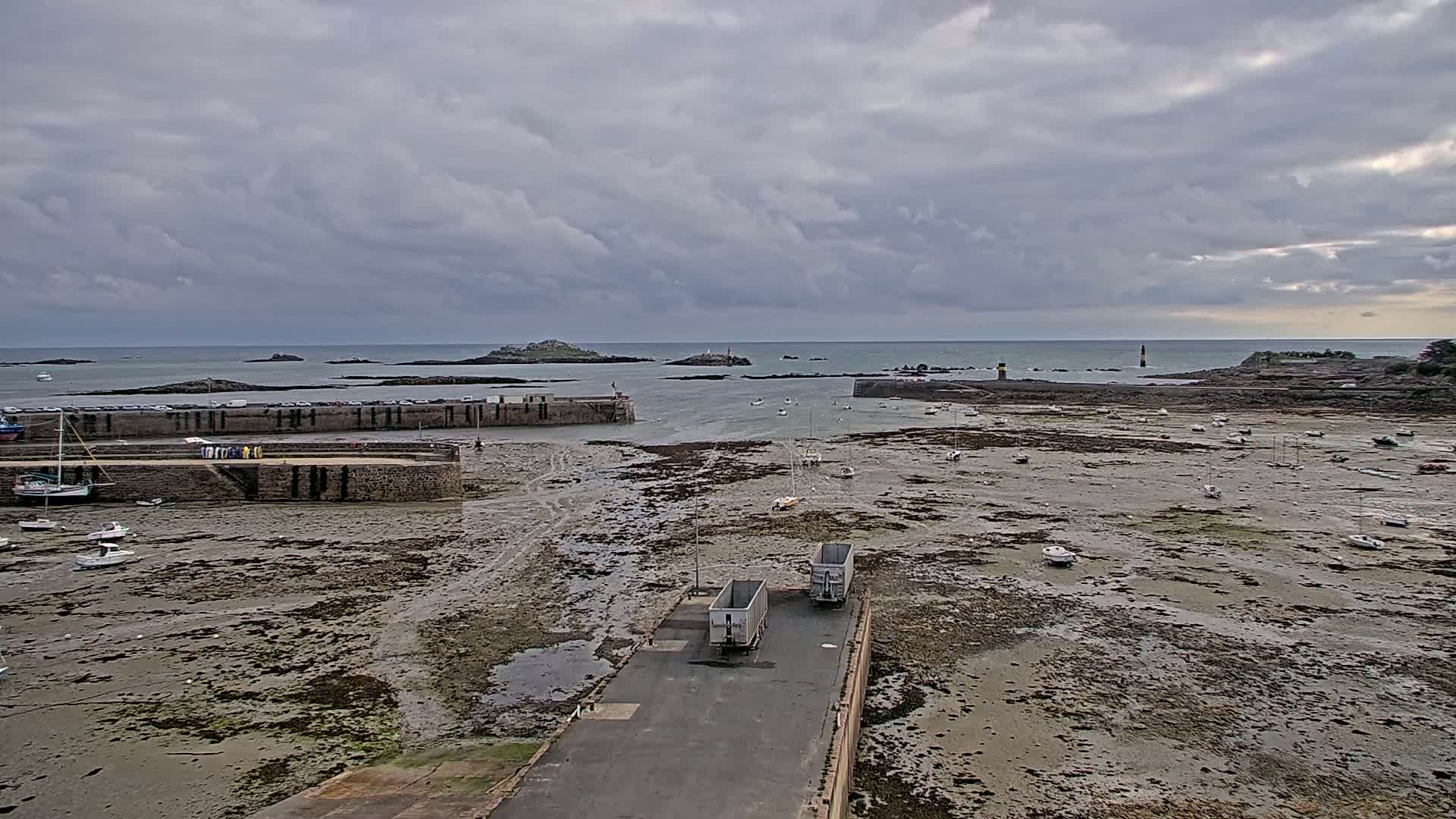 A harbor with numerous boats sits on exposed mudflats under a cloudy sky, with two cargo containers parked on a nearby road and islands visible in the distance.