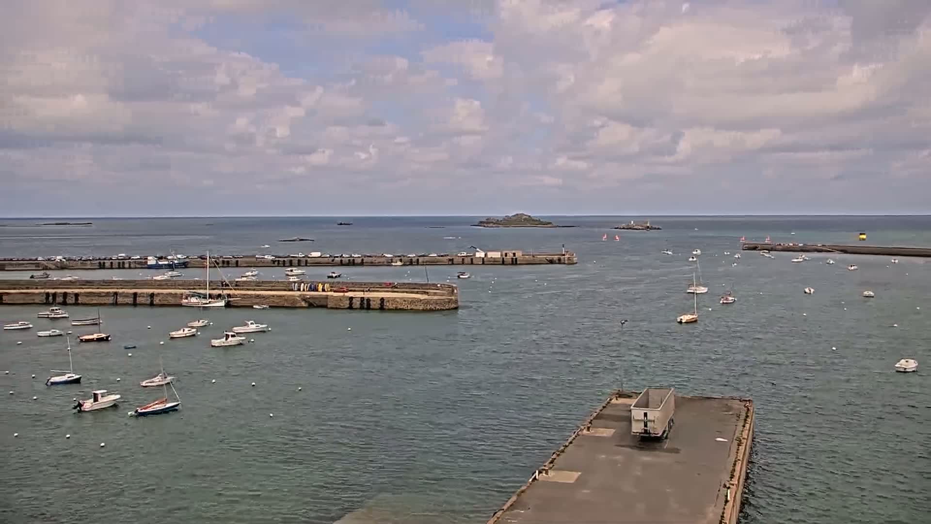 A harbor filled with numerous small boats under a partly cloudy sky, with a larger body of water and a distant island visible in the background.