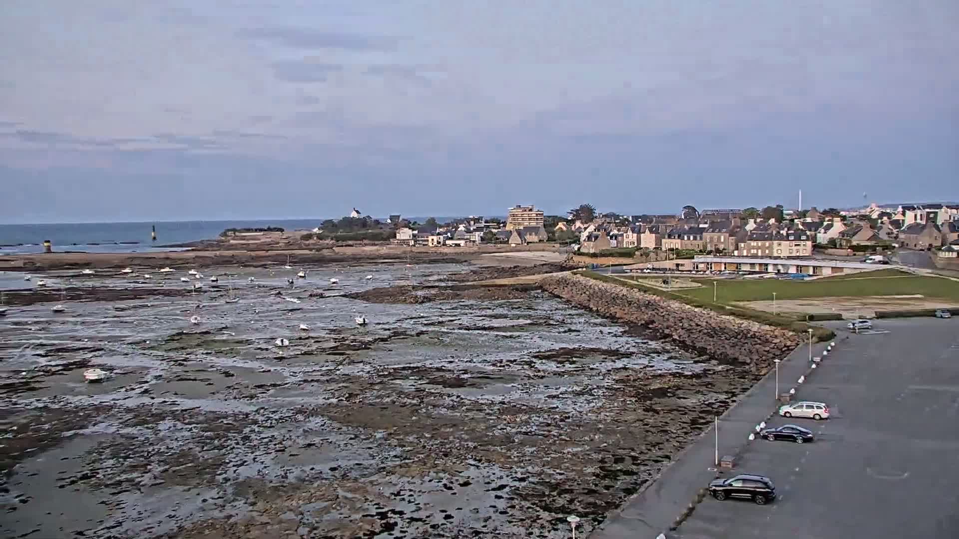 A coastal town overlooks a rocky, low-tide intertidal zone with numerous small boats scattered throughout, under a mostly cloudy sky.