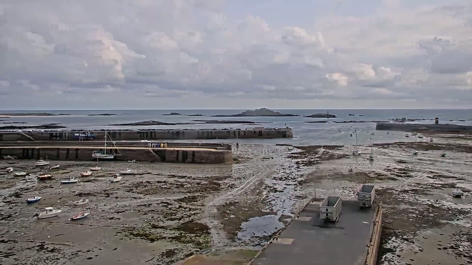 A harbor with numerous small boats sits mostly exposed at low tide under a cloudy sky, with two large trucks parked near the water's edge.