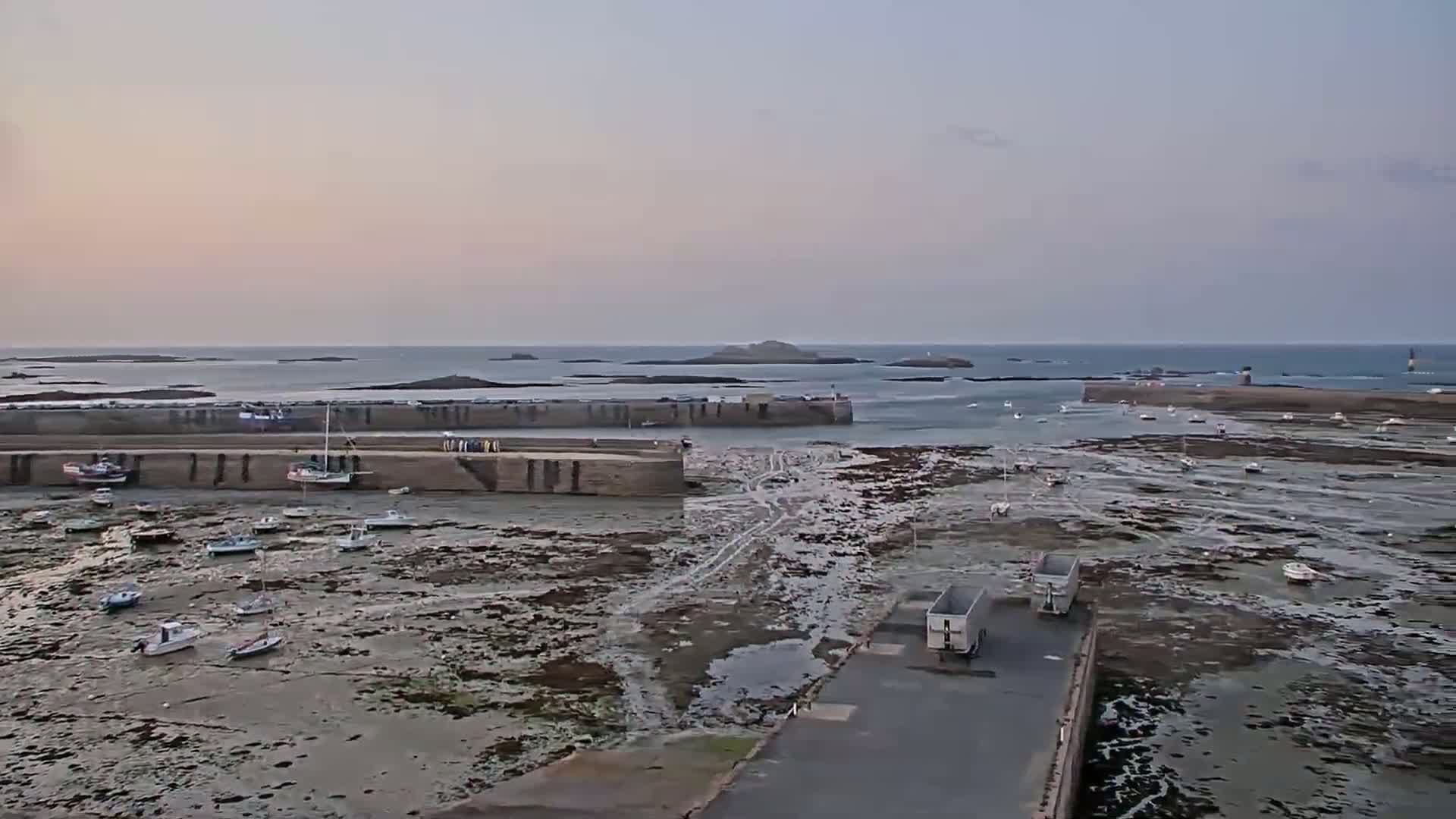 A harbor with many small boats sits on a mostly exposed seabed at low tide under a pale sunset sky.